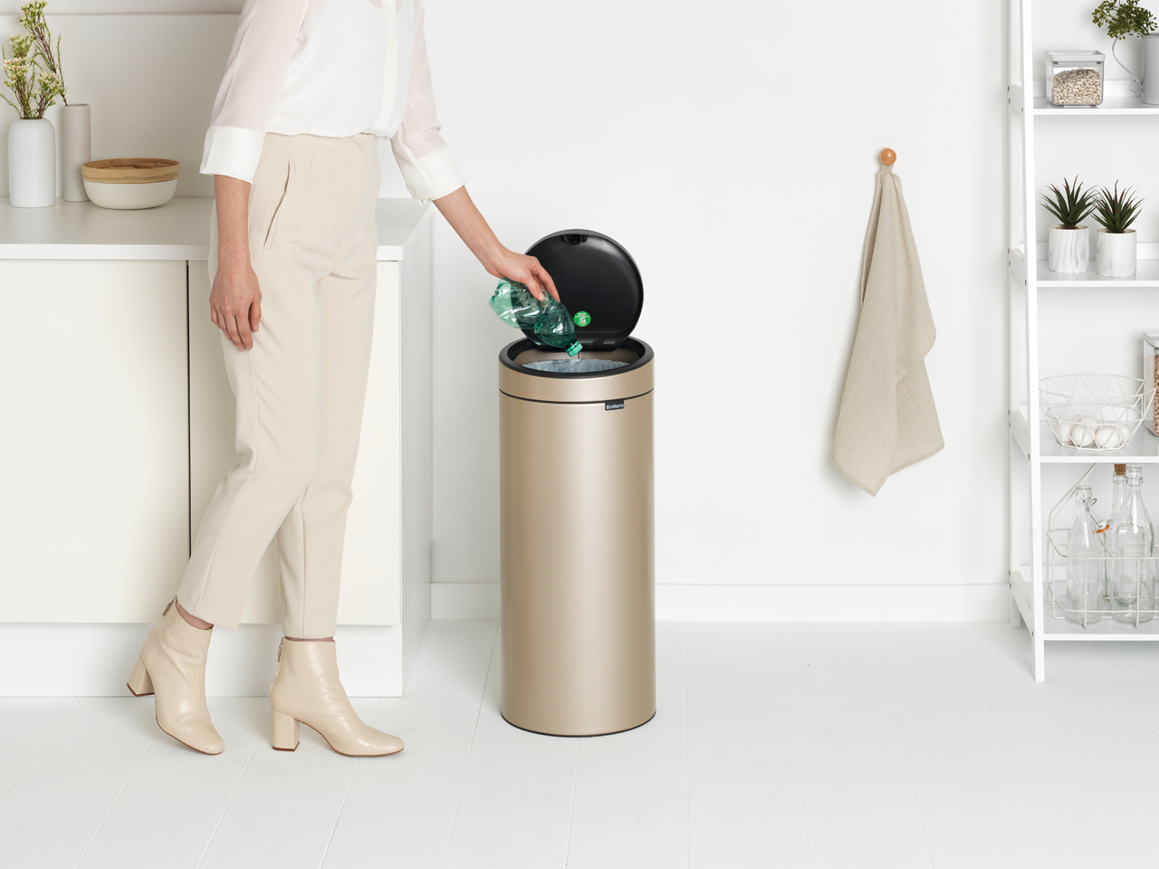 Eine Person wirft eine Plastikflasche in einen goldenen Mülleimer in einem hellen Raum. Weißer Hintergrund.