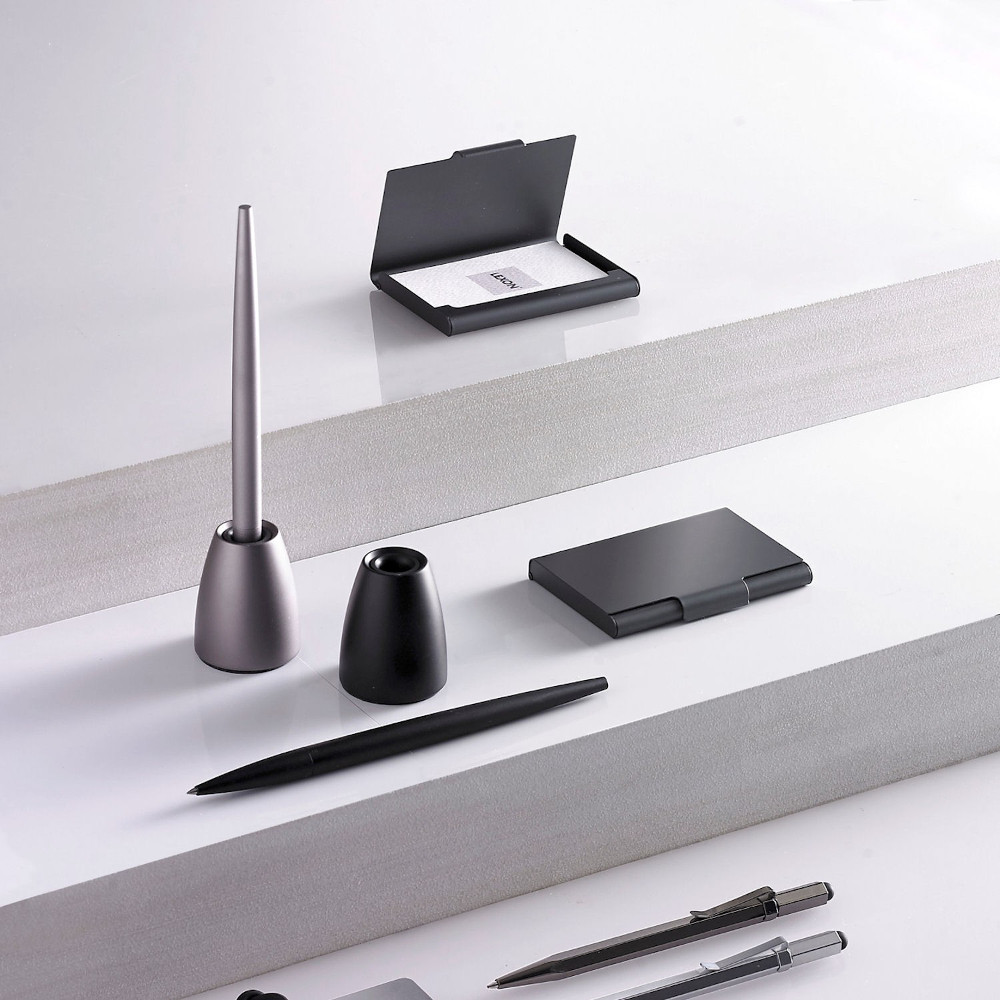 Artykuły biurowe na białych schodach. Ułożone są długopisy, piórniki i wizytownik. Przedmioty są czarne i srebrne.