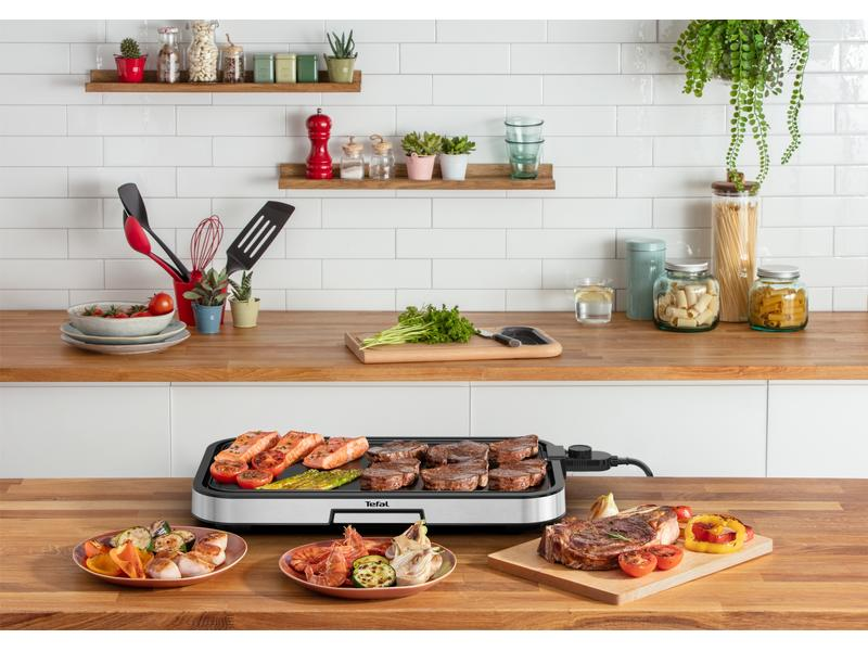 Elektrischer Grill mit Fleisch, Fisch und Garnelen, daneben Teller mit Beilagen.