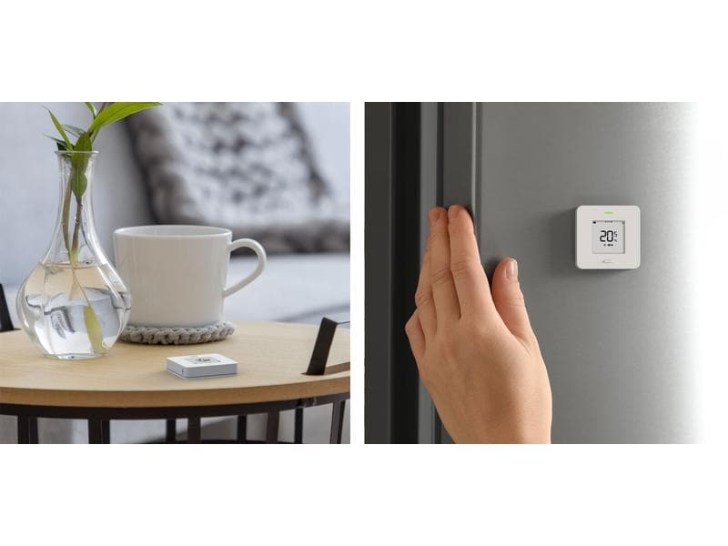 Un termostato intelligente bianco su un tavolo con vaso e tazza, e su una parete accanto a una mano.