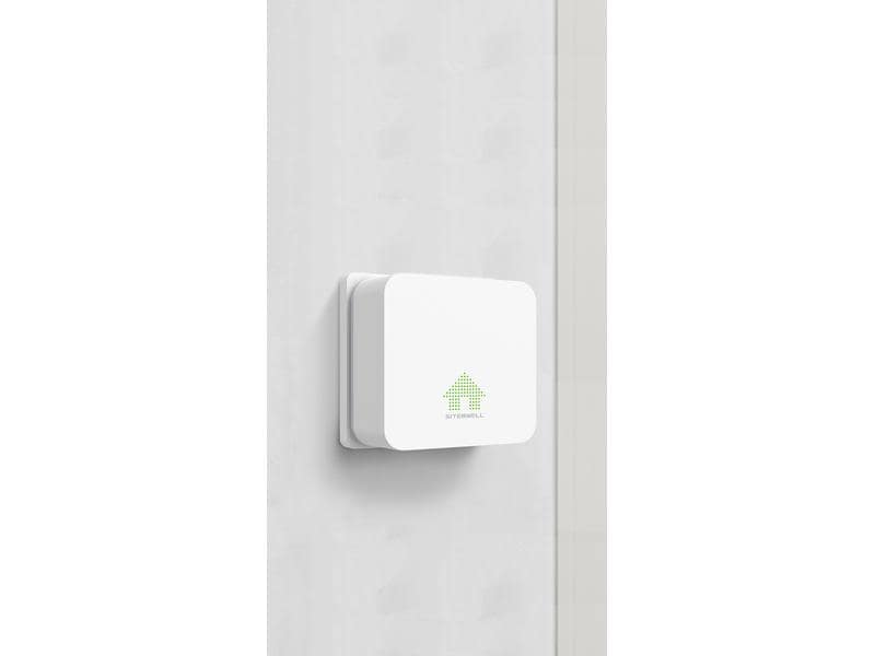 Ein weißes, quadratisches Smart-Home-Gerät mit grünem Haus-Symbol und Logo an einer Wand.