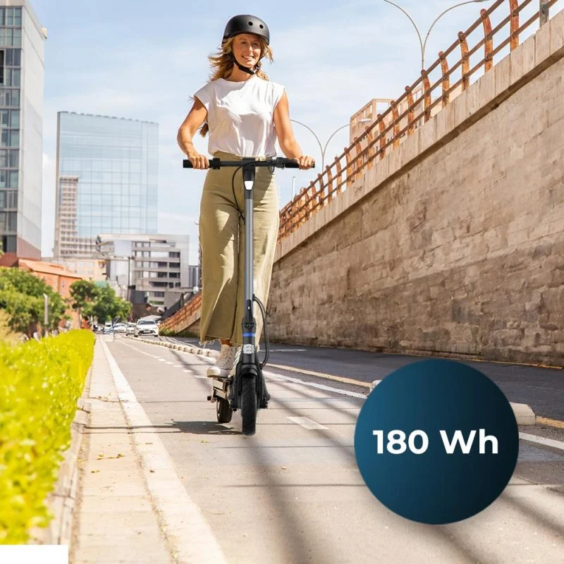 Kobieta jadąca skuterem elektrycznym po drodze. Budynki i most widoczne w tle.