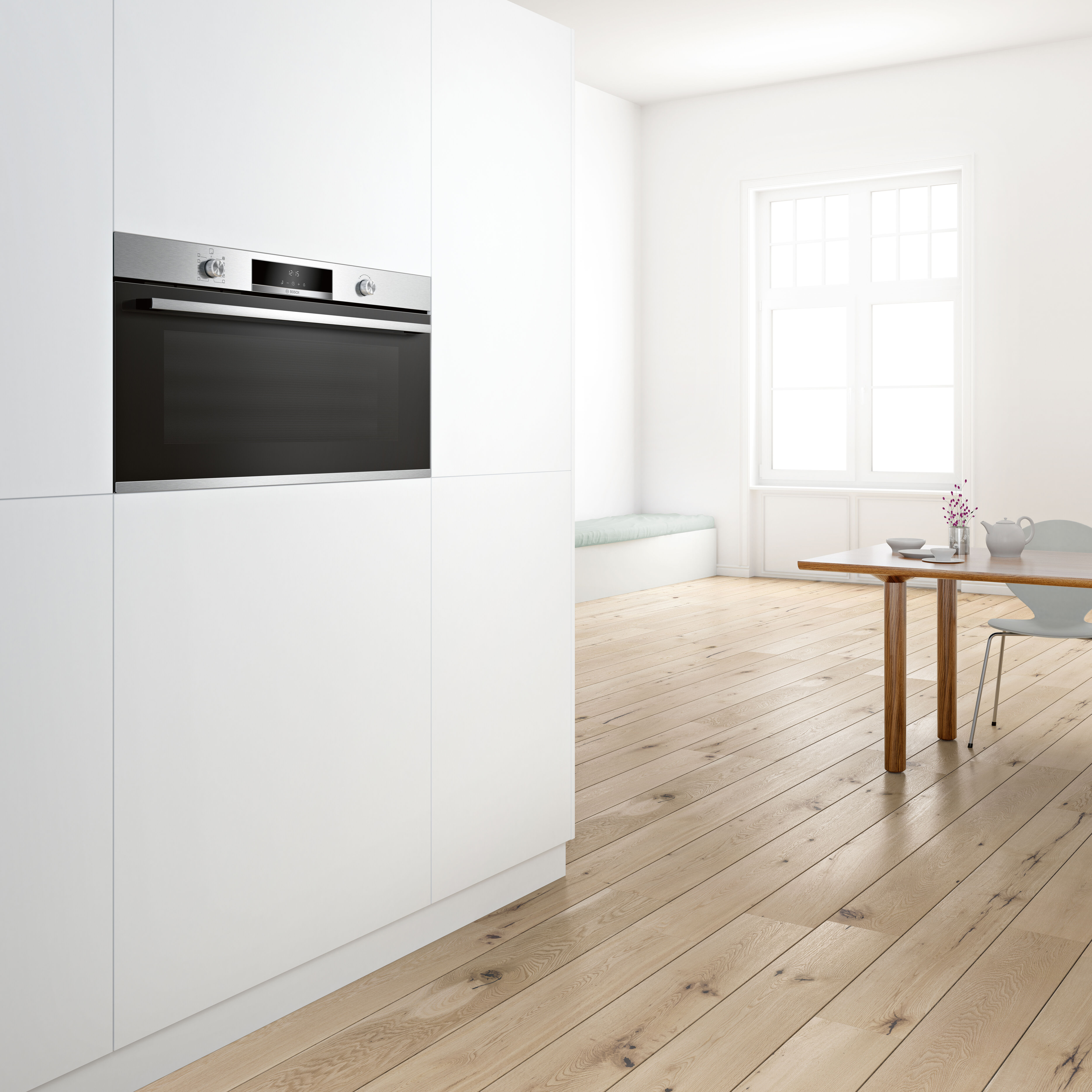 Cocina con armarios blancos, suelo de madera y horno de acero inoxidable. Mesa con platos al fondo.