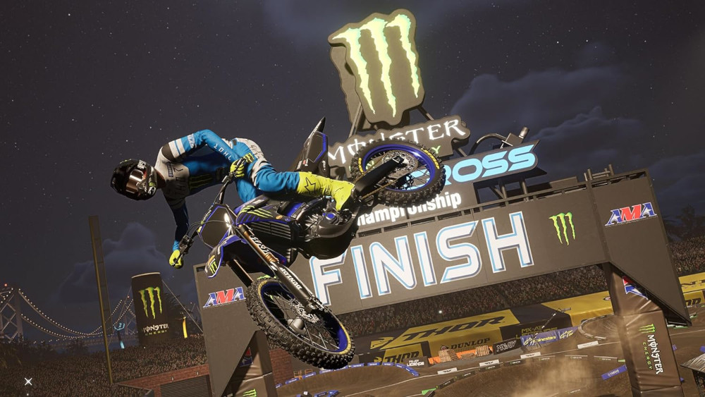 Motorcrosser in de lucht over een sprong, onder een 'Monster Energy'-banner 's nachts.