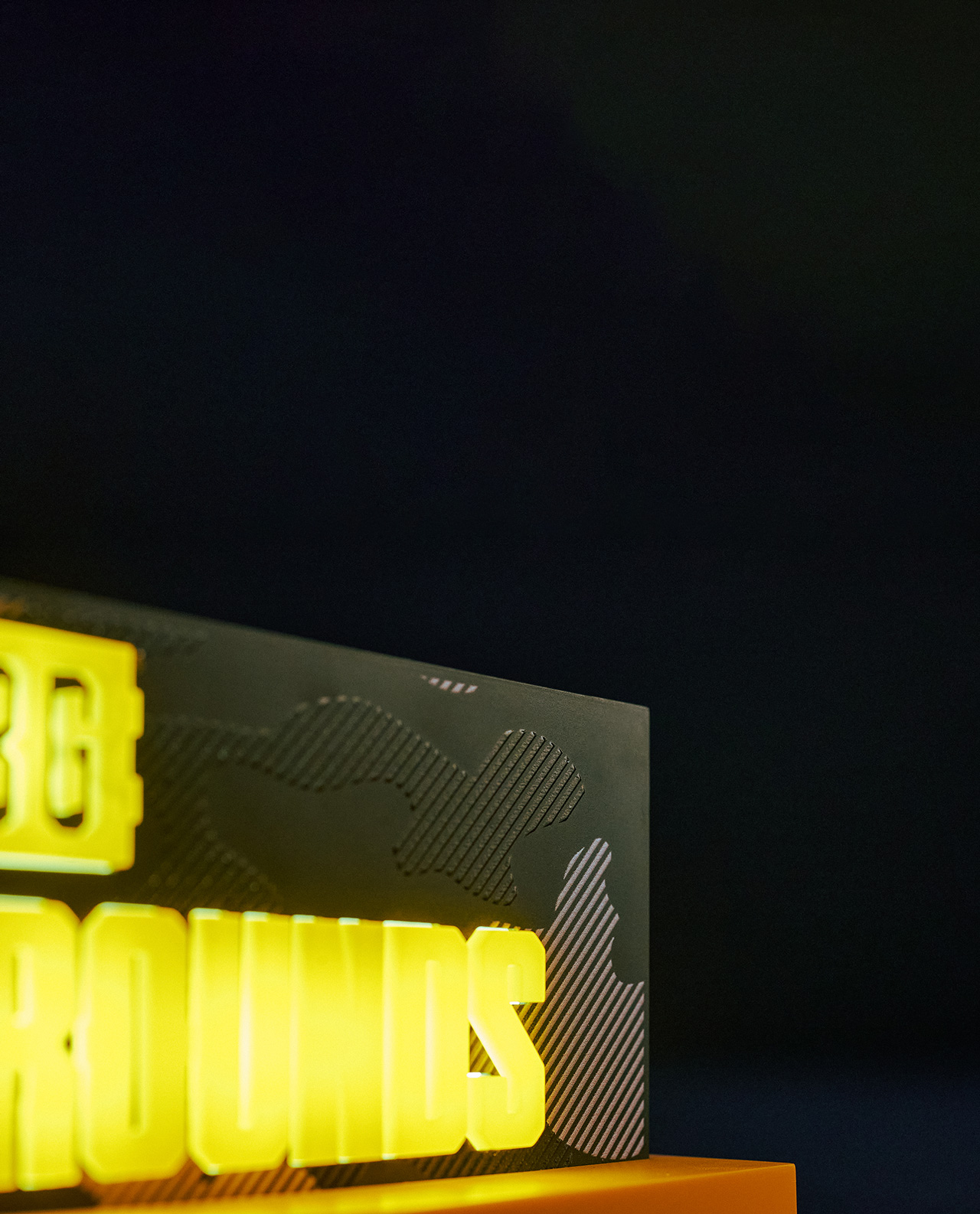 Beleuchtetes PUBG-Schild. Gelbe Schrift auf schwarzem und gemustertem Hintergrund. Oranger Sockel und schwarzer Hintergrund.
