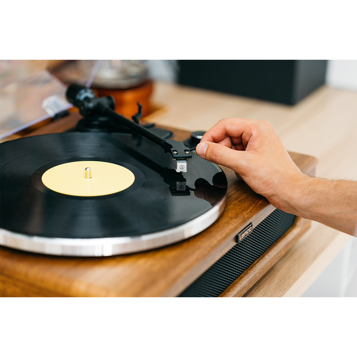 Una mano posiziona la puntina su un vinile su un giradischi in legno. Centro dorato.
