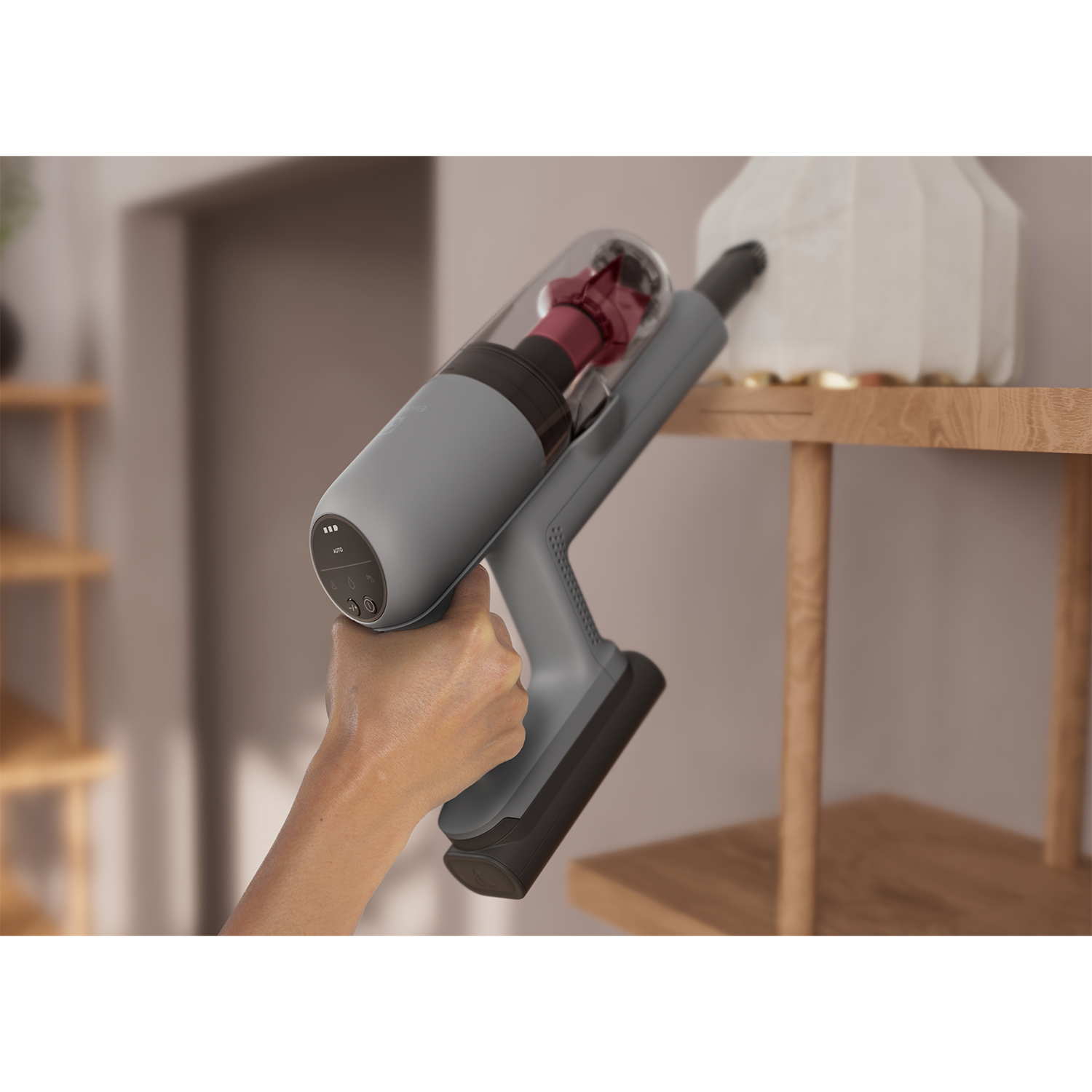 Aspirapolvere grigio, usato da una mano, accanto a una mensola di legno, su sfondo bianco.