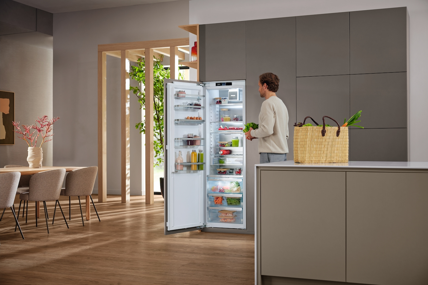 Mann stellt Lebensmittel in offenen Kühlschrank, daneben ein Korb mit Gemüse.