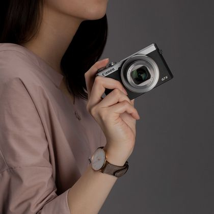Eine Frau hält eine Canon G7X Kamera, trägt eine Uhr. Grauer Hintergrund.