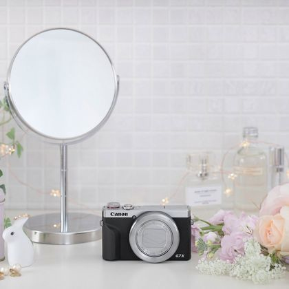 Silber-schwarze Kamera auf weißem Tisch, neben Spiegel, Blumen und Zubehör.