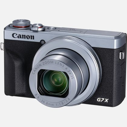 Eine silberne und schwarze Canon G7X Kamera auf weißer Oberfläche. Das Objektiv ist sichtbar.