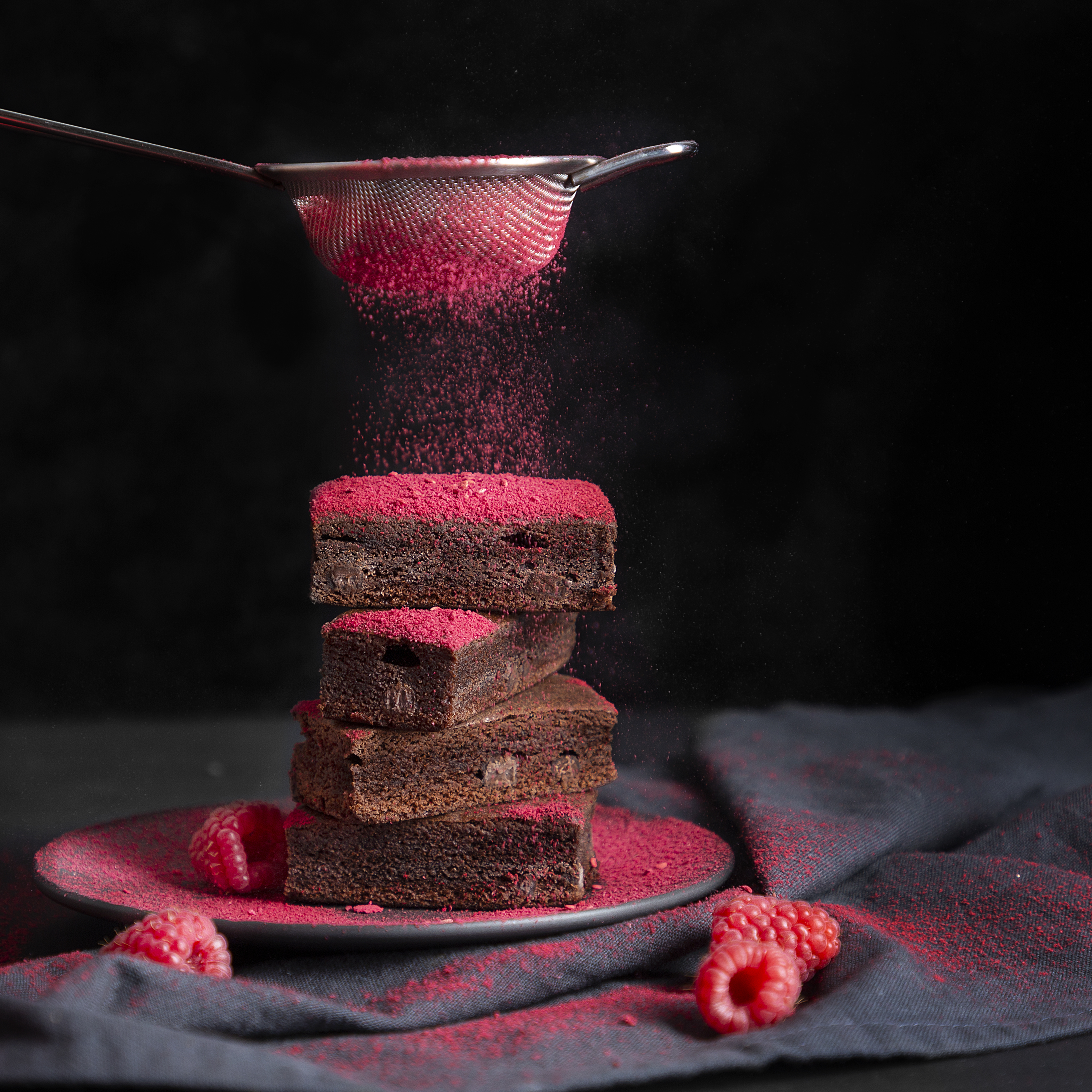 Ein Stapel Brownies wird mit rosa Puder bestäubt. Himbeeren sind auf einem Teller und auf der Oberfläche.