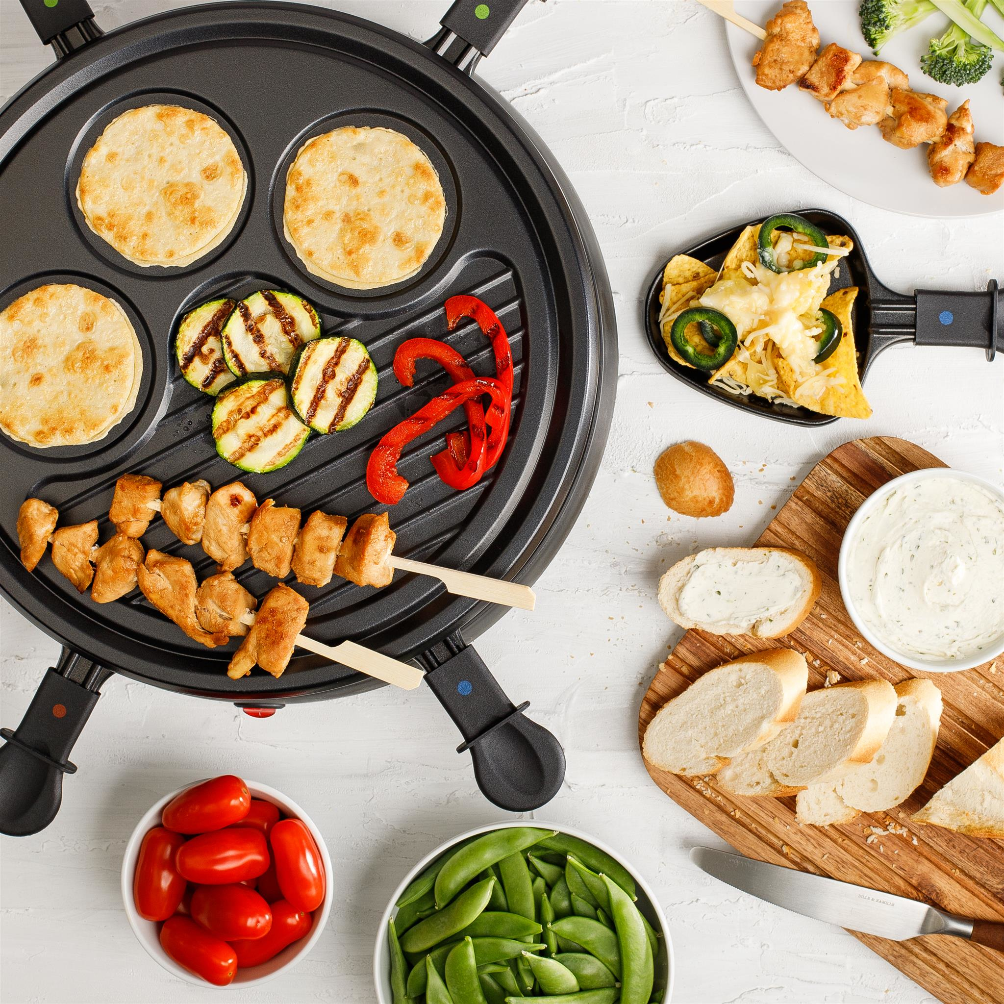 Draufsicht auf einen Raclette-Grill mit gekochten Speisen: Pfannkuchen, gegrilltes Gemüse, Spieße, Nachos, Brot, Tomaten und Erbsen.