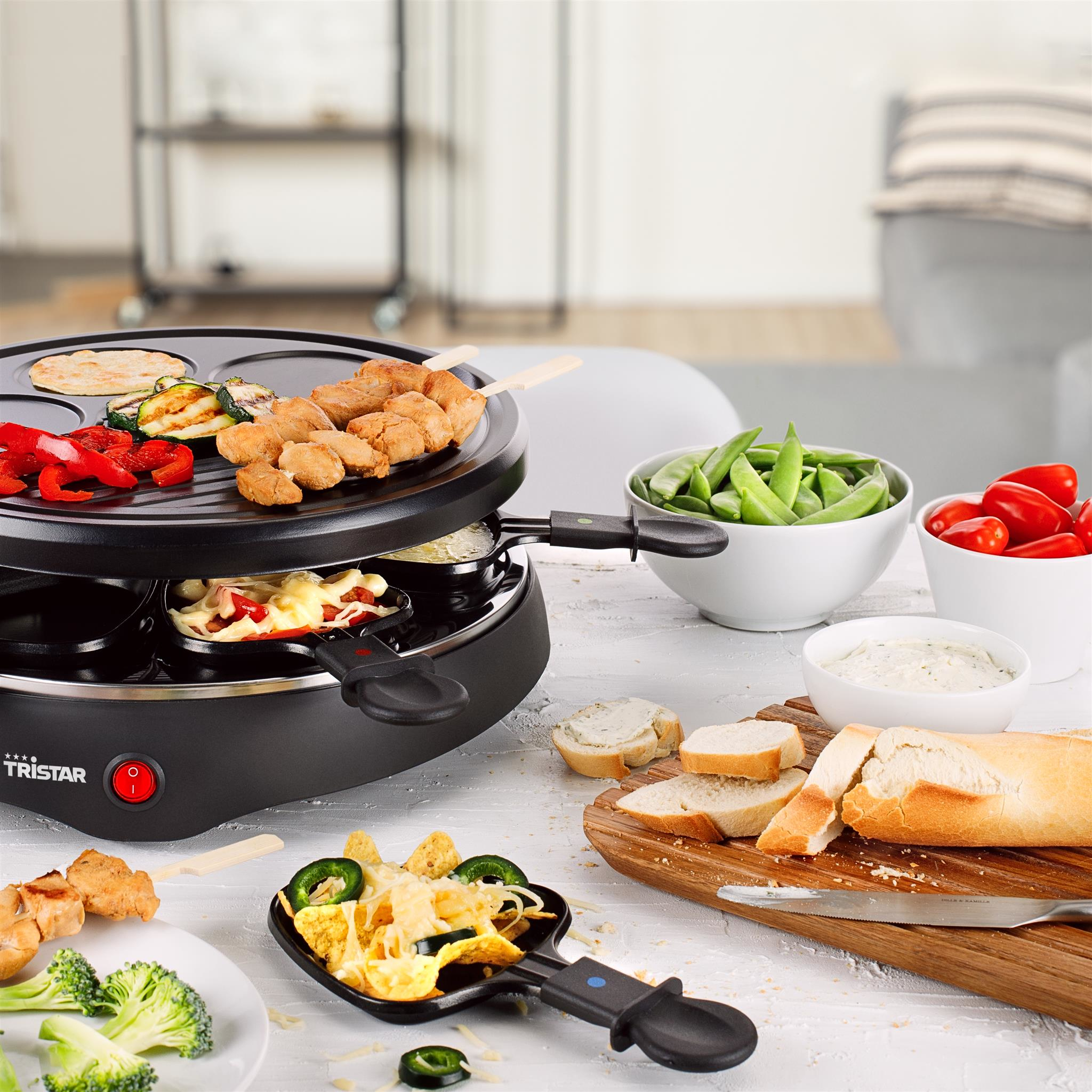 Raclette-Grill mit gekochten Speisen. Gemüse, Käse und Fleisch sind abgebildet. Brot, Dips und Beilagen sind ebenfalls vorhanden.