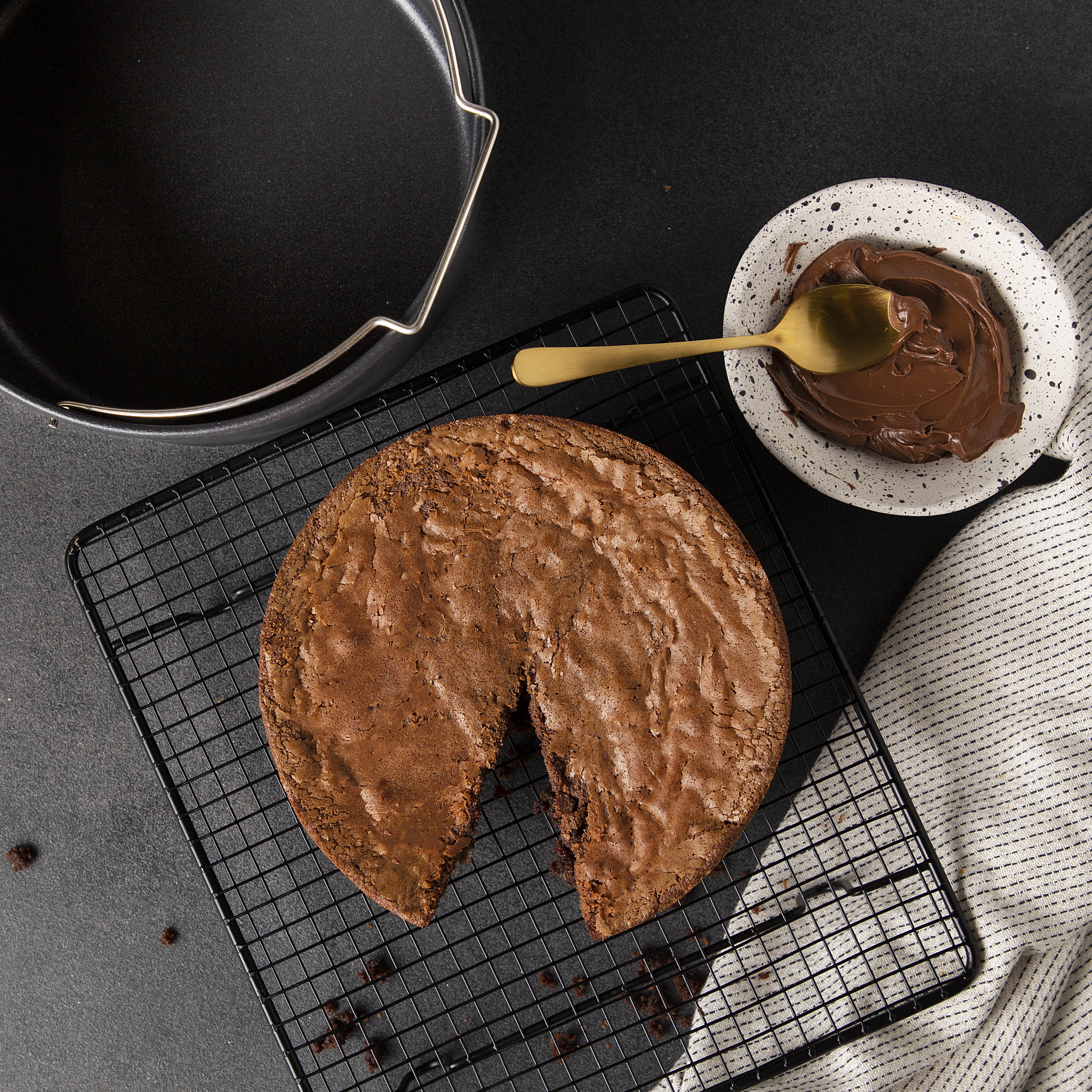 Chocolate cake on a wire rack, cut slice missing, and bowl of melted chocolate.