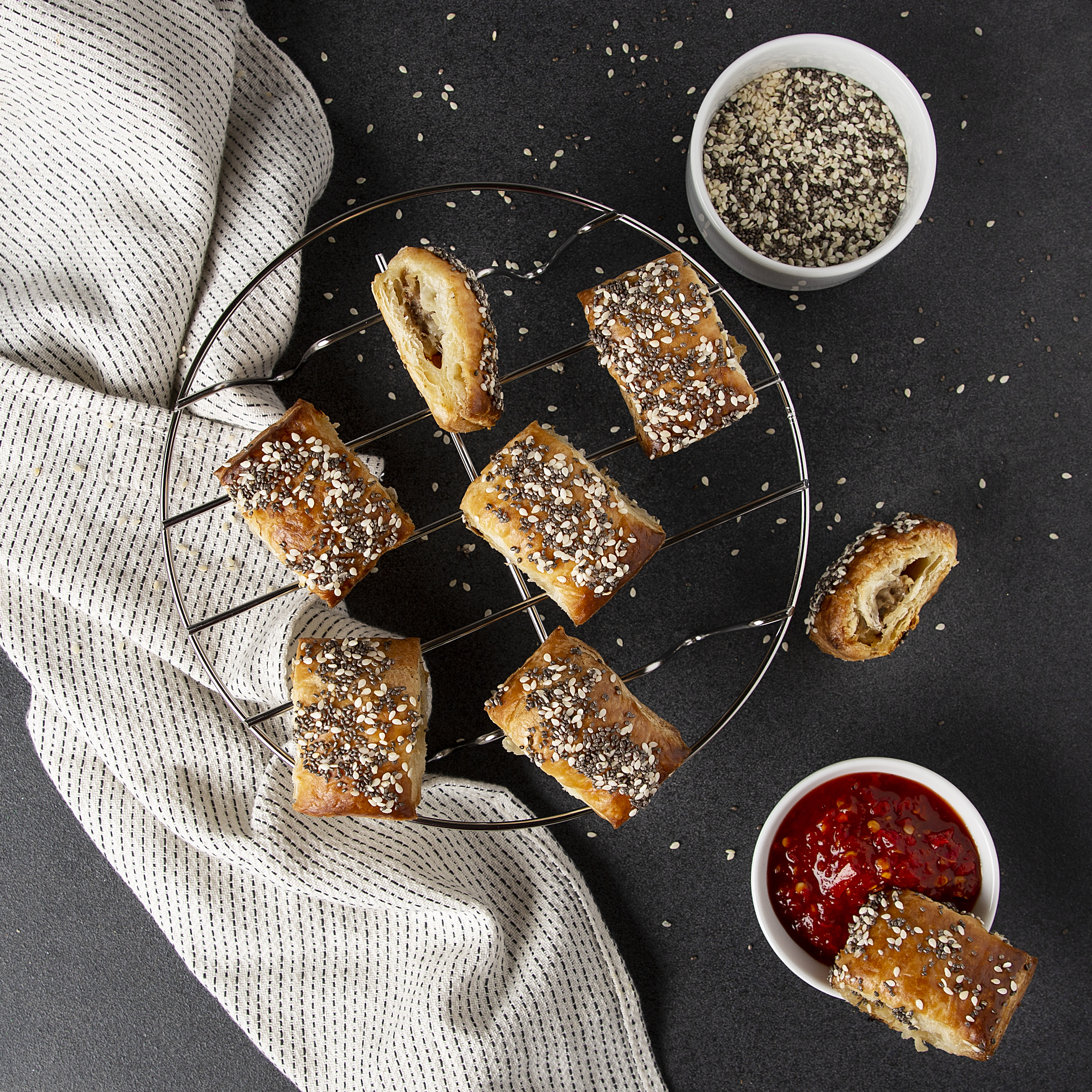 Overhead shot of pastry rolls with seeds, a cooling rack, and two bowls with seeds and sauce.