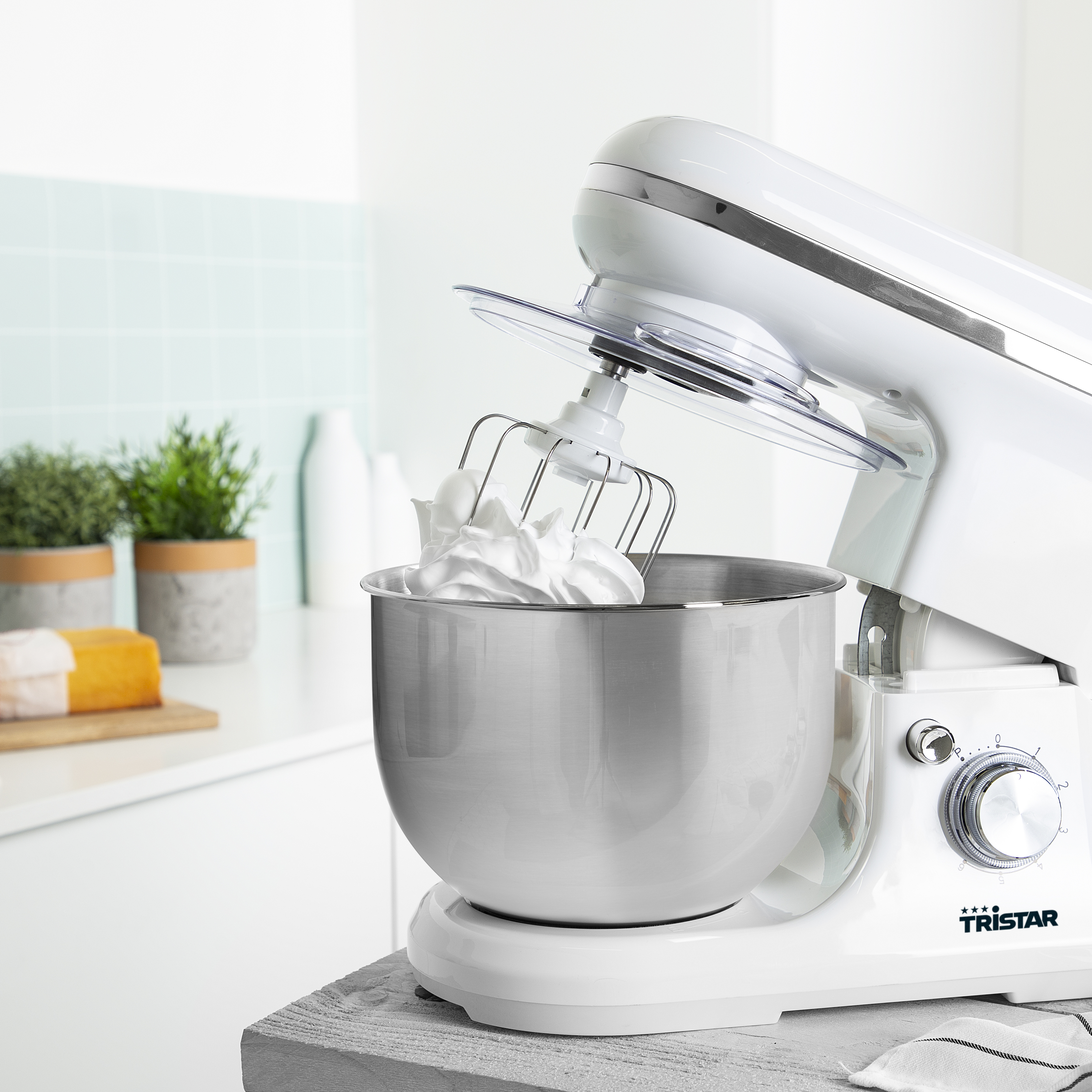Robot culinaire blanc avec bol, fouet et substance blanche. L'arrière-plan comprend une cuisine et des plantes vertes.