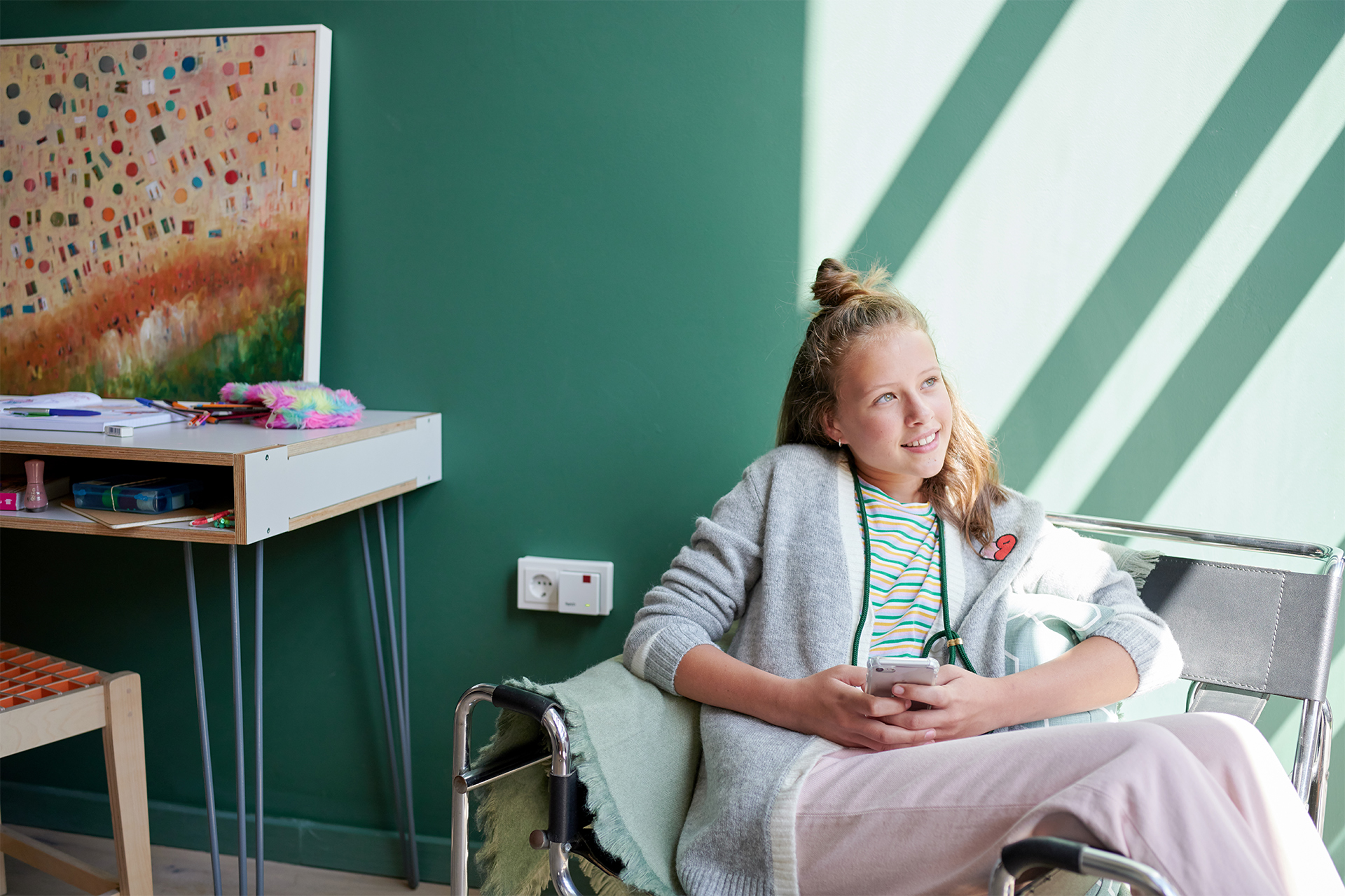 Ein Mädchen sitzt mit Handy, trägt Pullover, Streifenhemd, rosa Hose. Grüner Wand im Hintergrund.