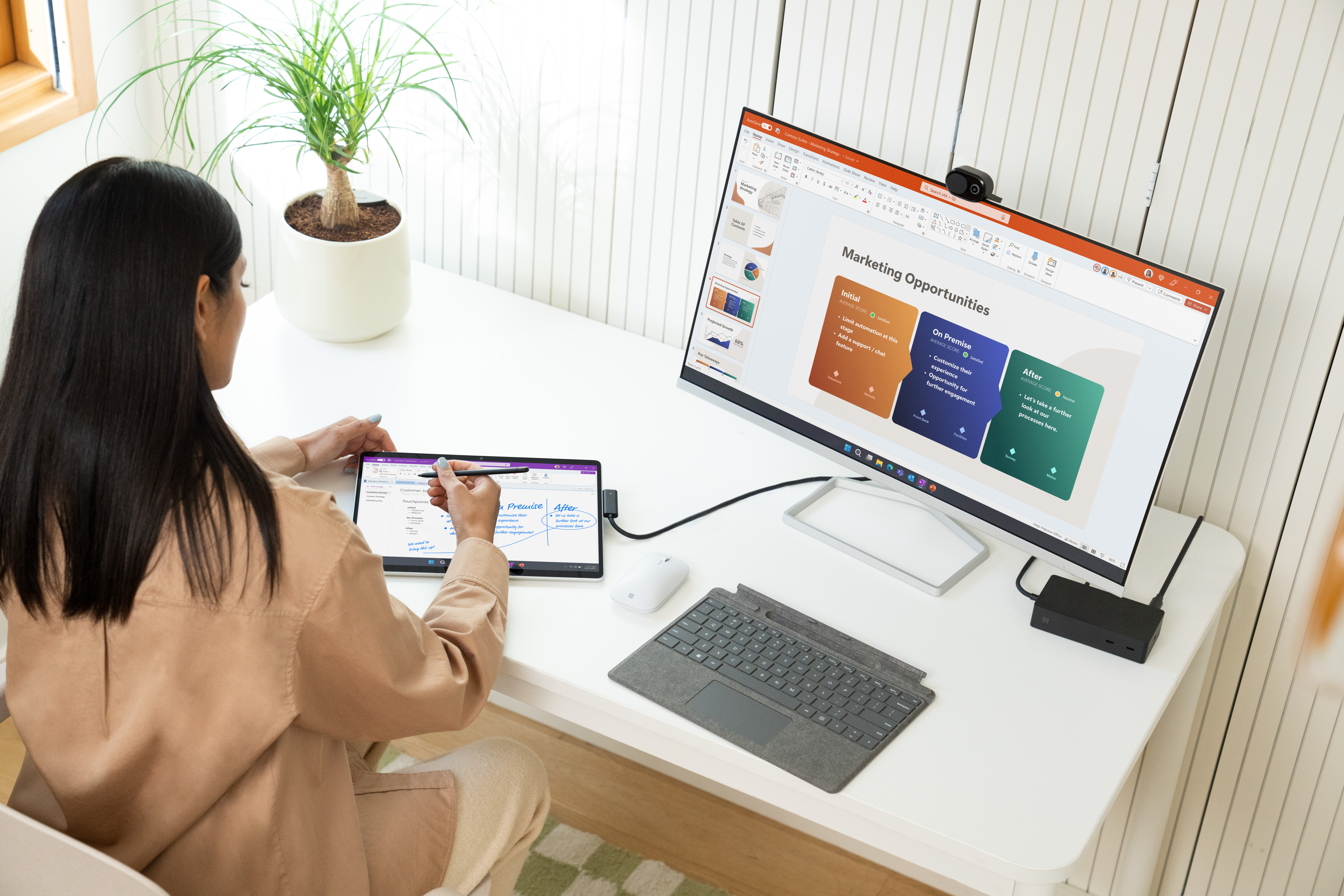 Vrouw gebruikt tablet en monitor op wit bureau. Plant links.