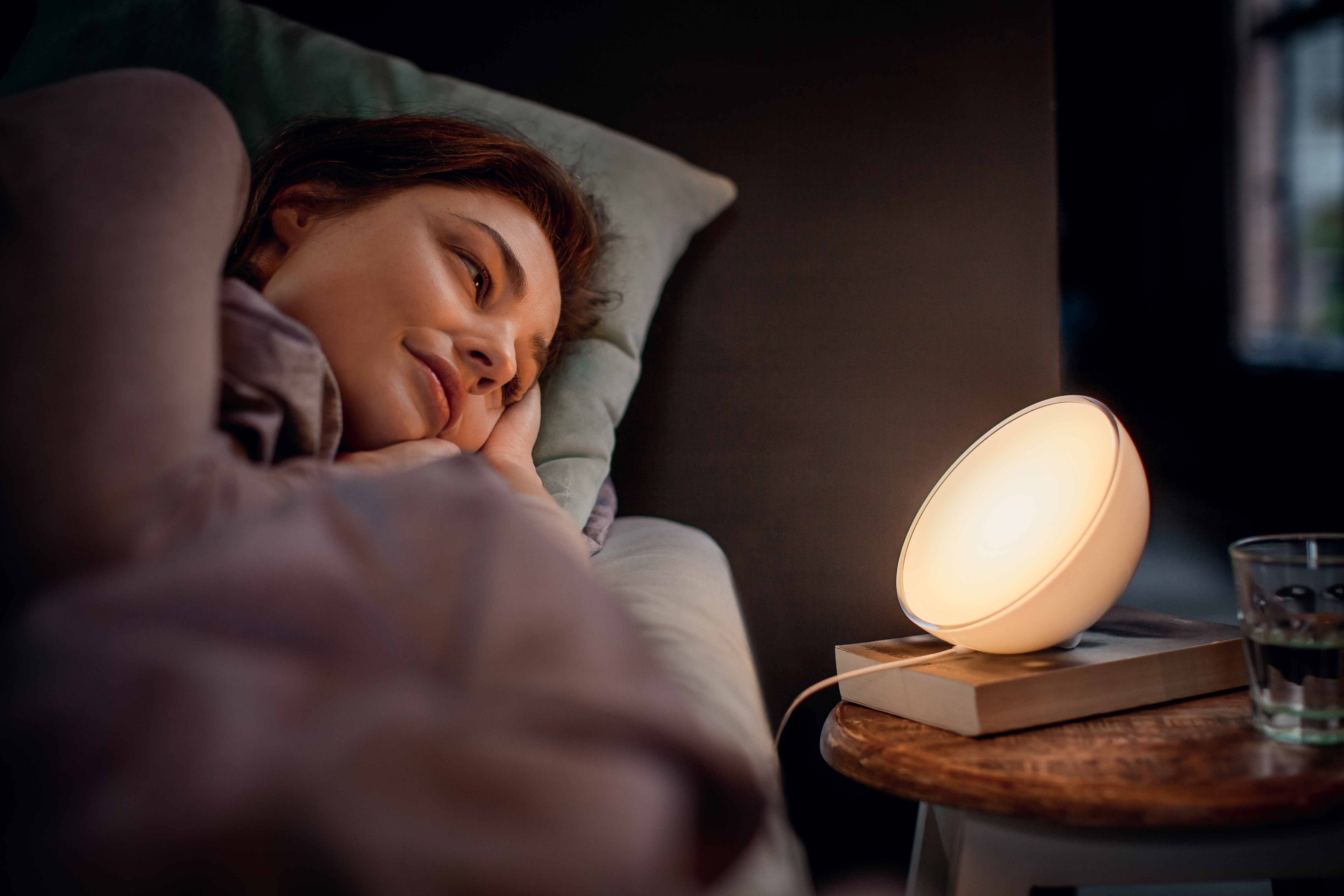 Frau im Bett, blickt auf eine leuchtende Nachttischlampe mit Buch und Wasserglas.
