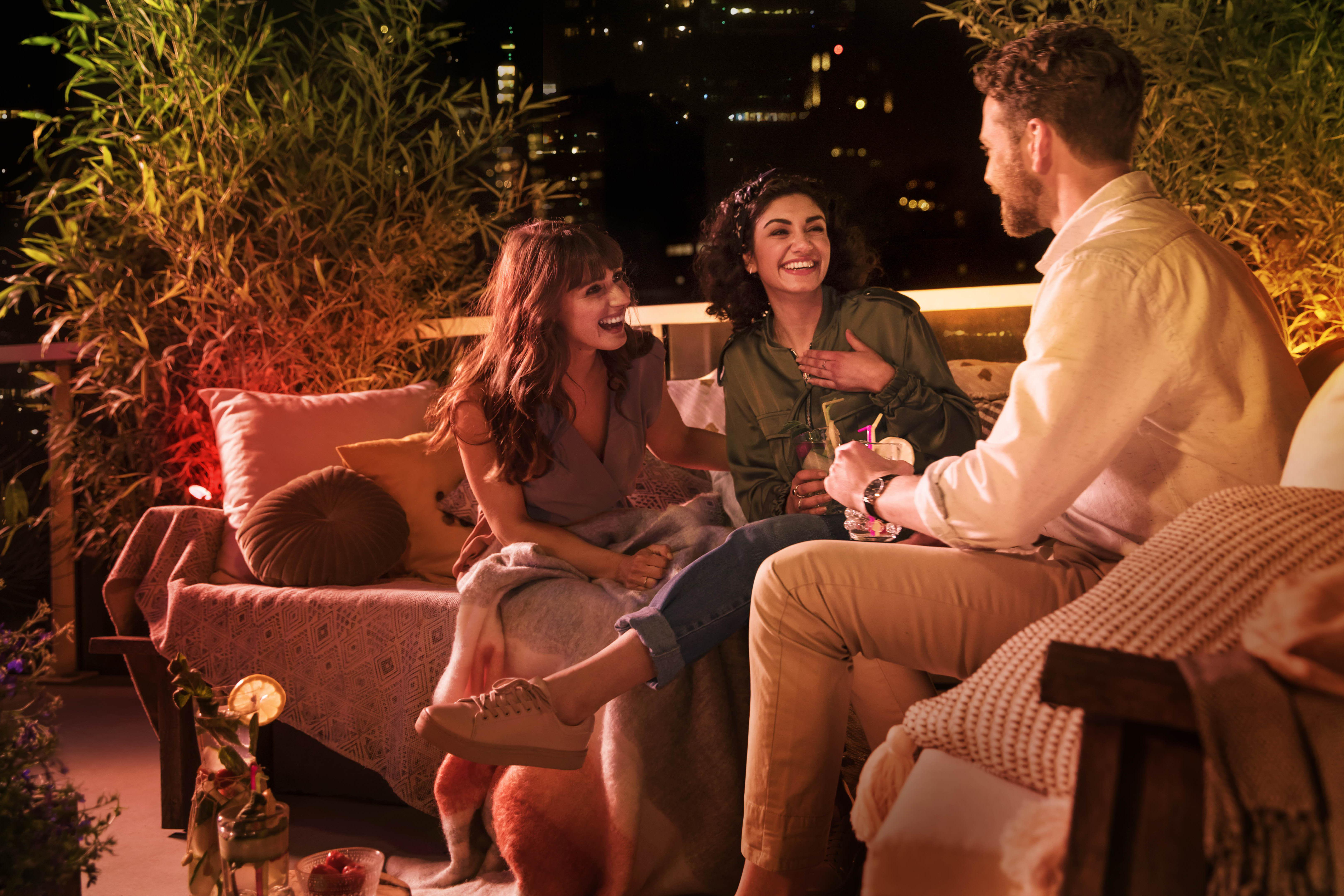 Drei Personen sitzen abends auf einer Terrasse, mit sanfter Beleuchtung und Getränken.