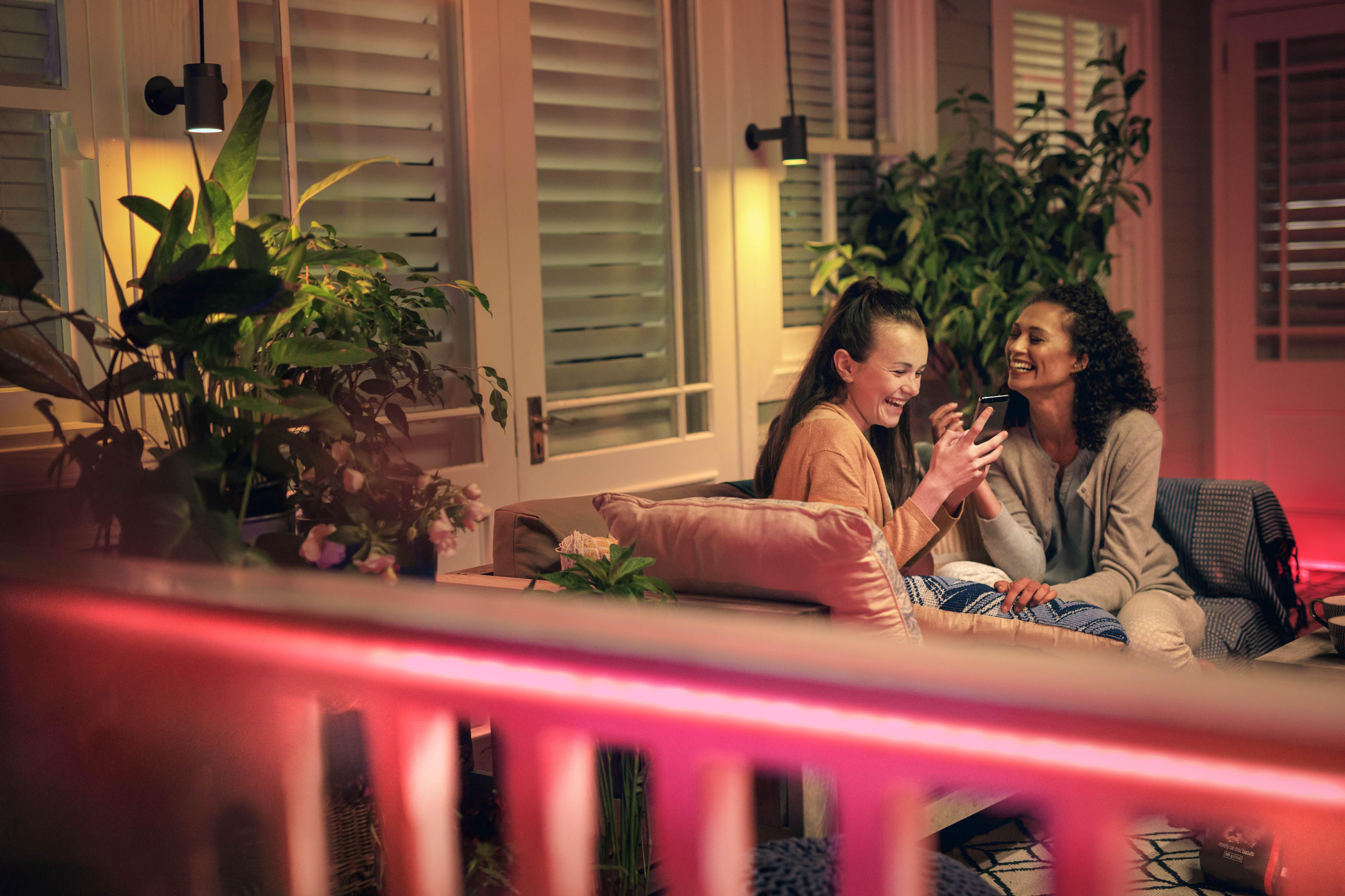 Zwei Frauen auf einer Veranda, benutzen Telefone. Pflanzen und Lichter sind sichtbar. Rosa und gelbe Lichter sind an.