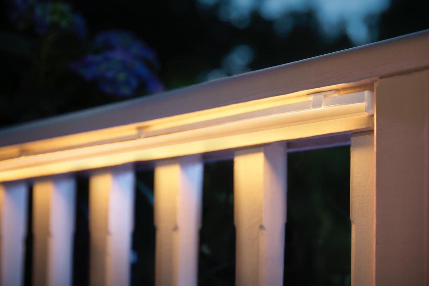 Weißes Holzgeländer mit beleuchtetem Lichtband. Blaue Blumen sind im Hintergrund leicht sichtbar.
