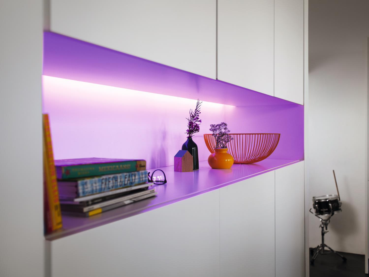 Ein weißes Regal mit lila LED-Licht mit Büchern, Brille und orangefarbener Vase. Schwarze Trommel im Hintergrund.