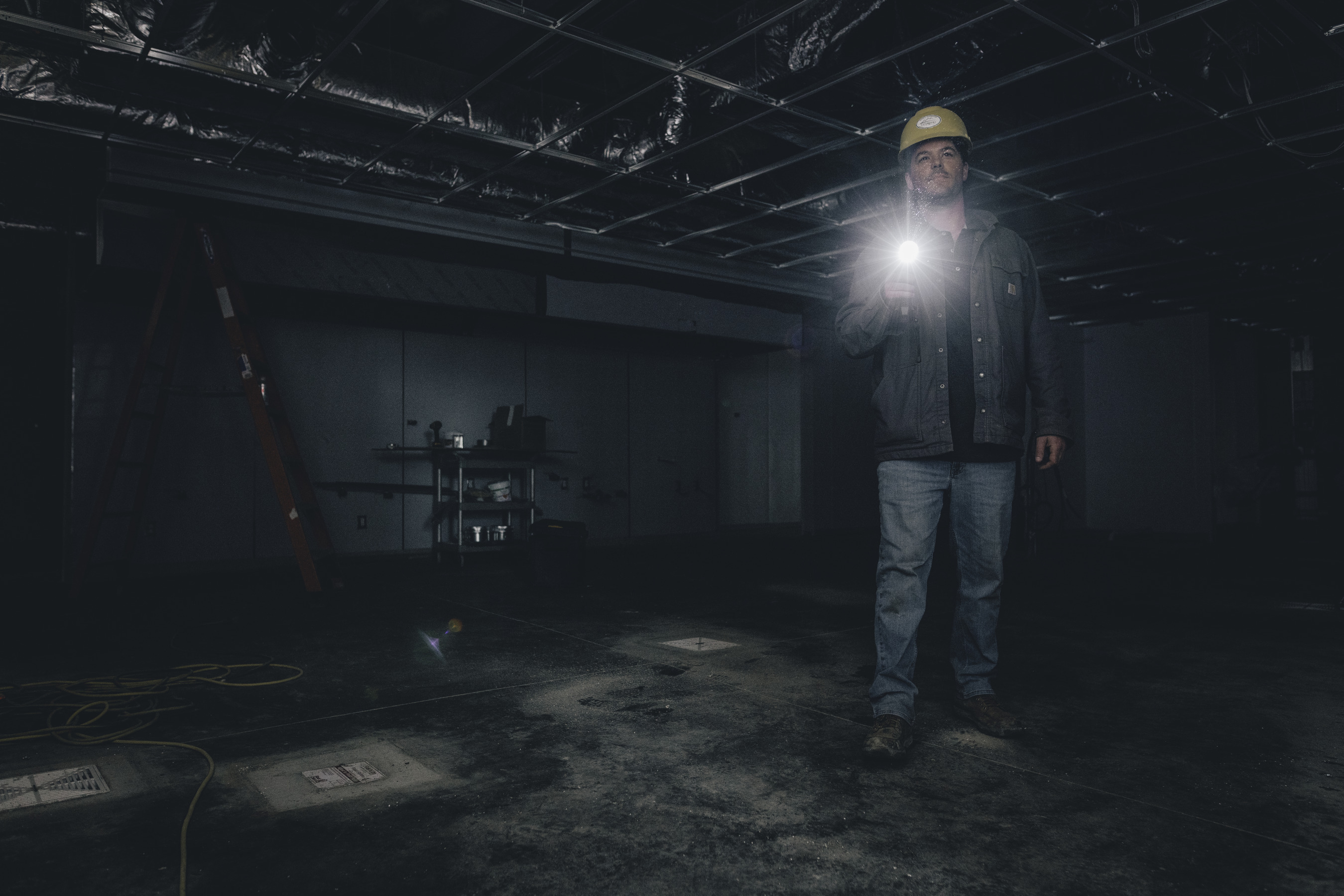 Bauarbeiter mit Taschenlampe in dunklem Raum, Leiter und Rohre im Hintergrund.