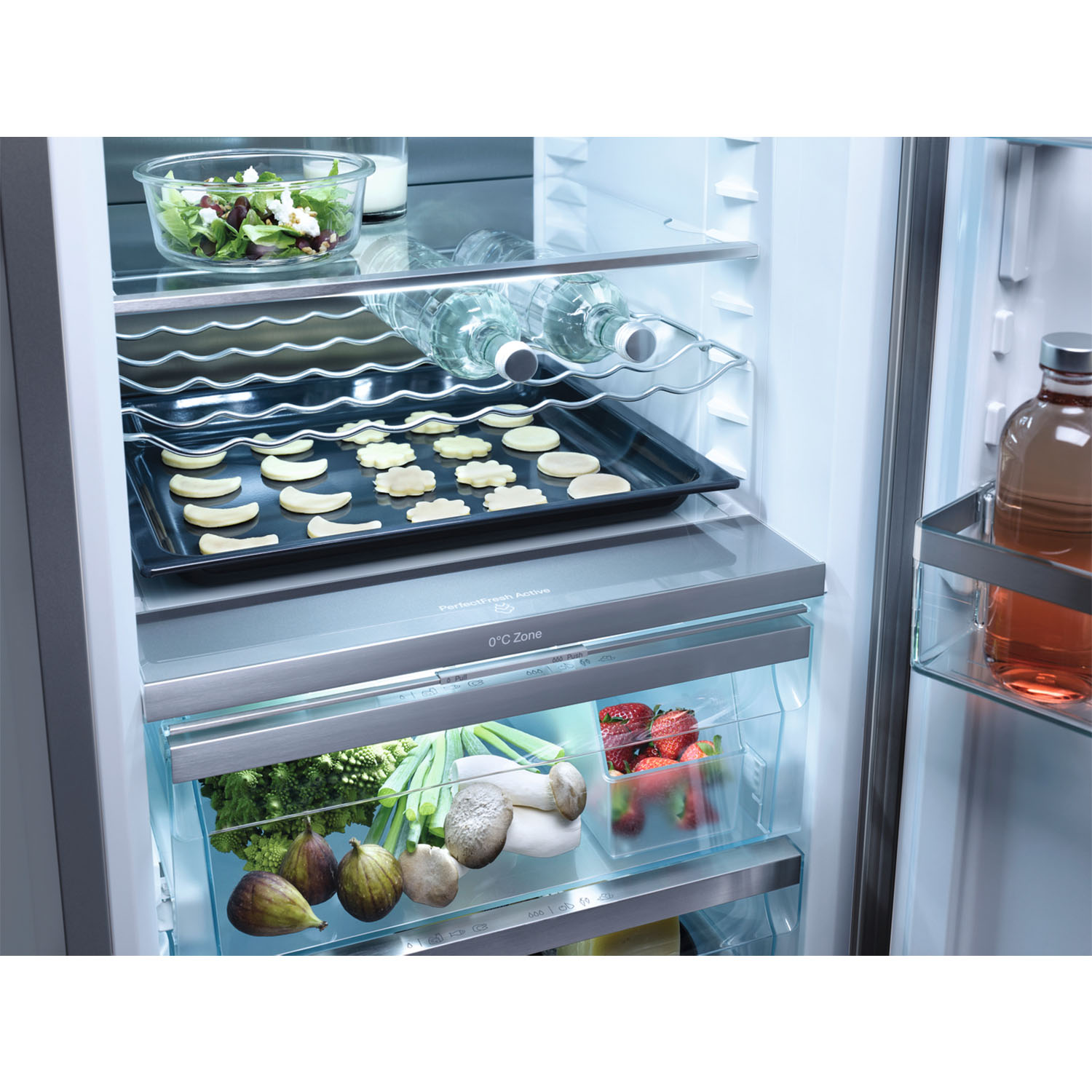 Frigorifero aperto con alimenti come insalata, bottiglie d'acqua, vassoio di biscotti e prodotti freschi.