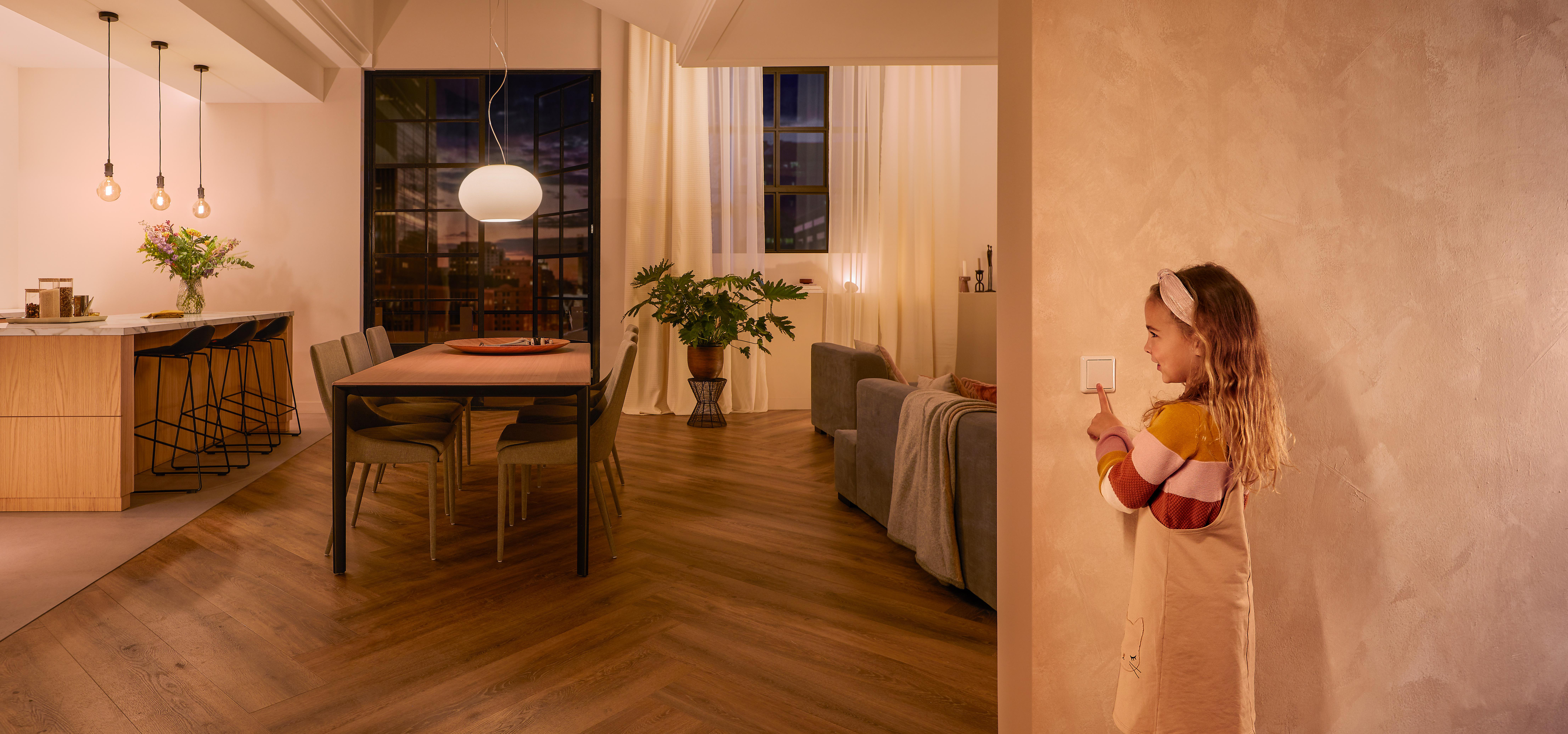 Mädchen berührt Lichtschalter in einem Raum. Warme Beleuchtung, modernes Interieur, Essbereich im Hintergrund.