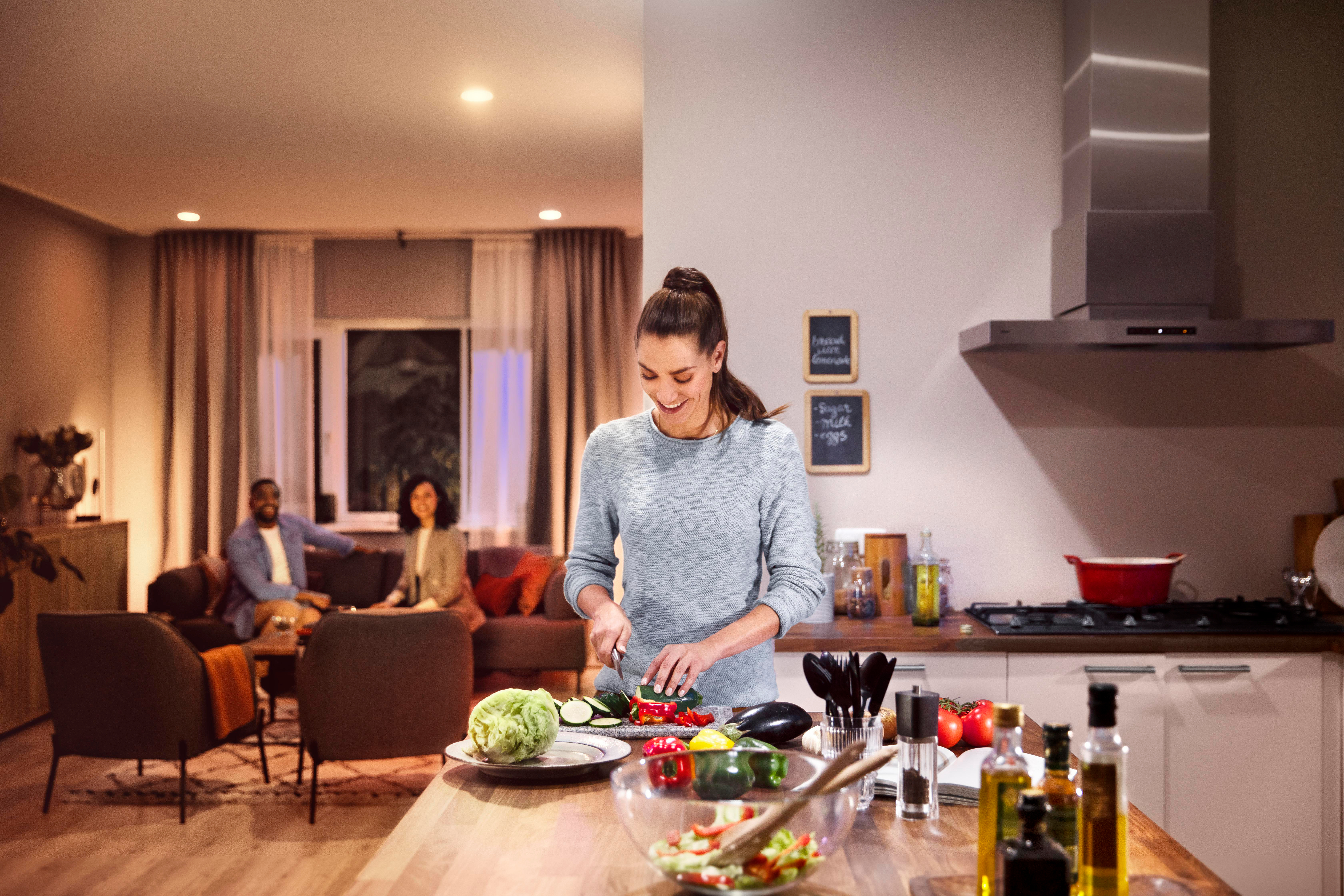 Frau schneidet Gemüse in gut beleuchteter Küche, Personen im Hintergrund.