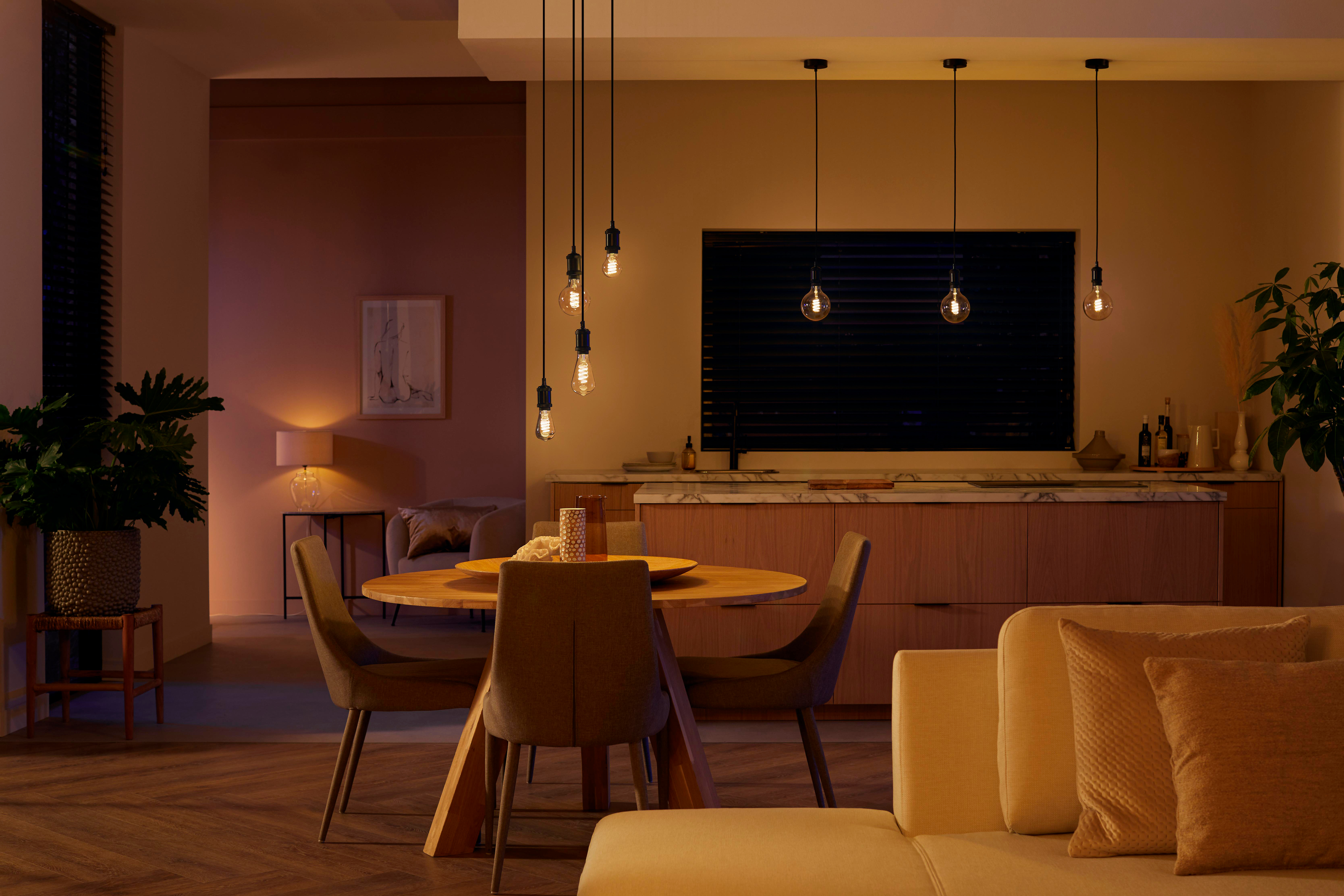 Wohnzimmer mit moderner Beleuchtung. Es sind Esstisch und Stühle, Sofa und ein großes Fenster zu sehen.