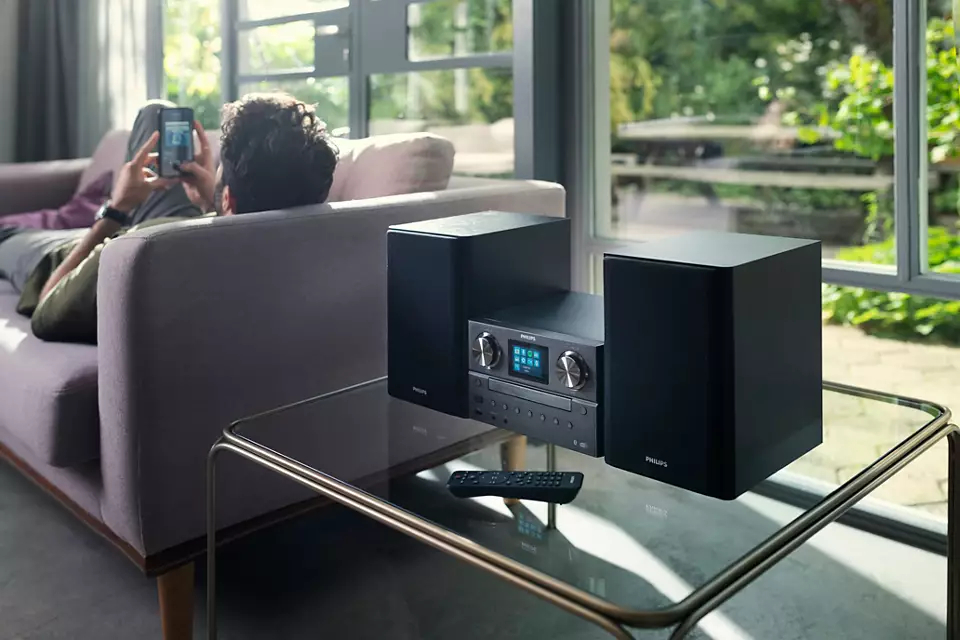 Mężczyzna na kanapie z telefonem, wieża stereo na stole i głośniki.