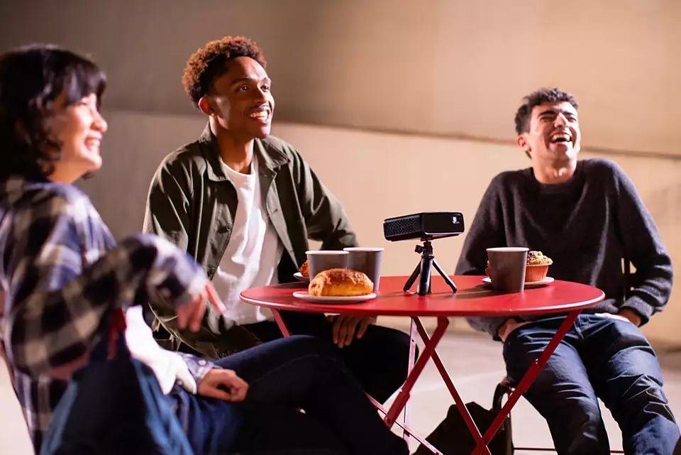 Trzy osoby śmieją się wokół czerwonego stołu z jedzeniem i projektorem.