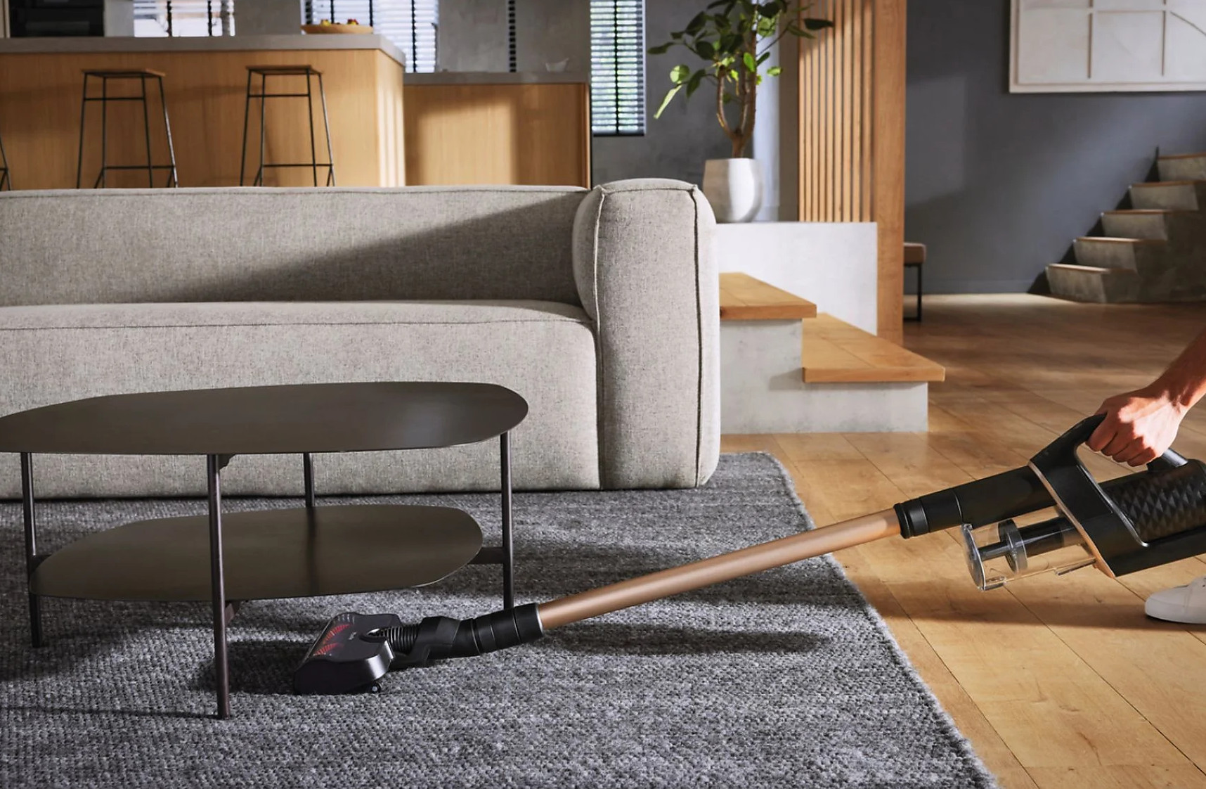 Osoba odkurzająca dywan odkurzaczem bezprzewodowym. Nowoczesny salon z szarą sofą.