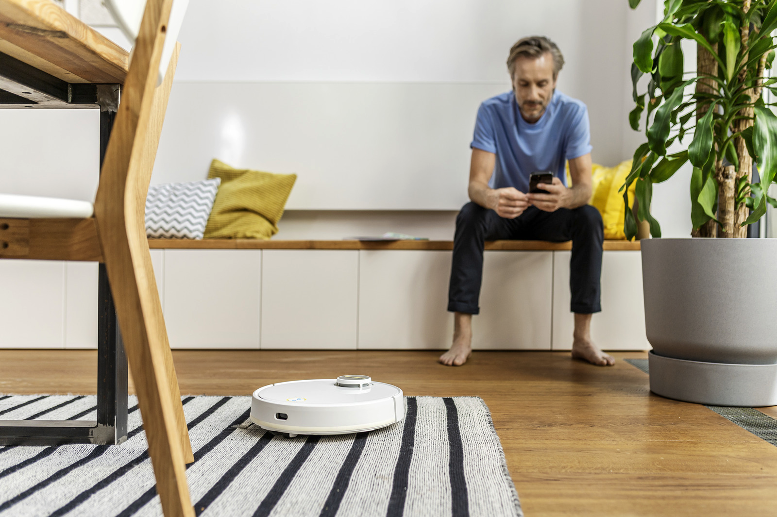 Egy férfi egy padon ülve a telefonját nézi, miközben egy robotporszívó takarítja a padlót.