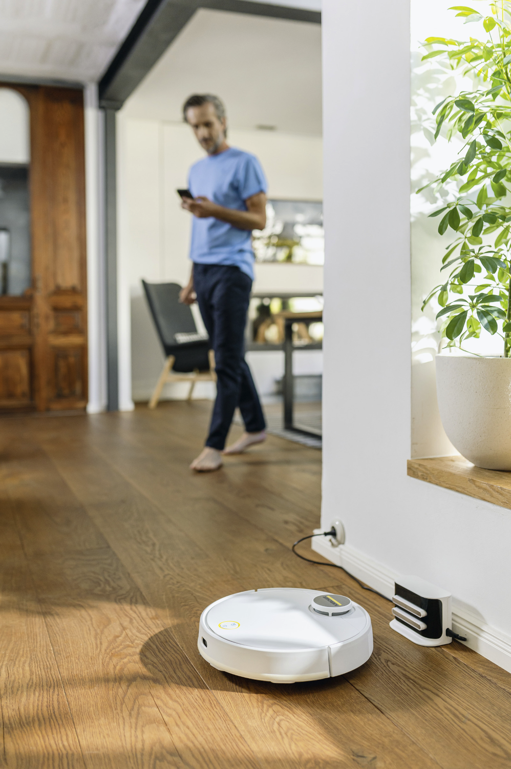 Robot porszívó a parkettán, kék inges férfi sétál a háttérben.
