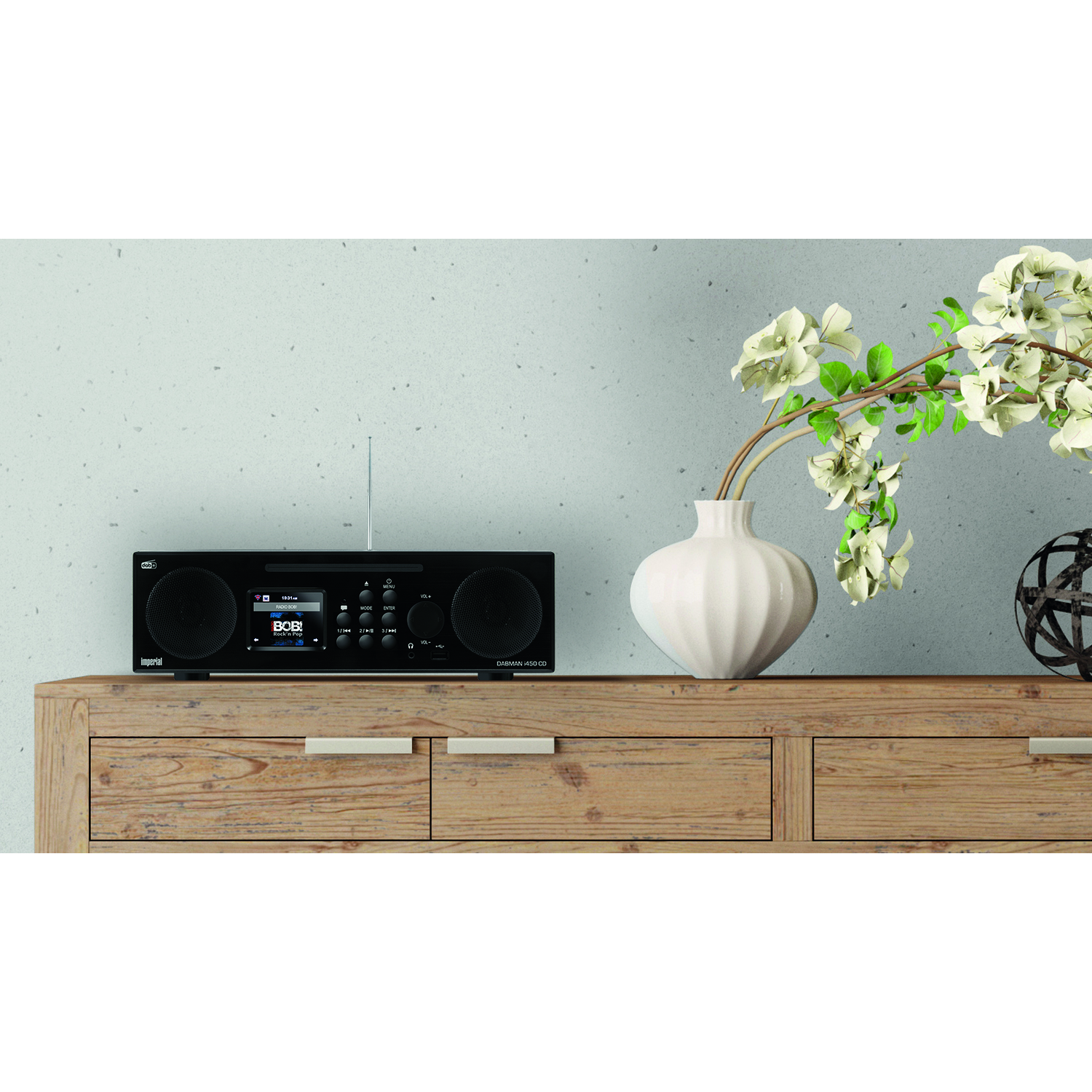 A black radio sits on a wooden cabinet, with a white vase of flowers to the right. A dark metal sphere sits next to the vase.