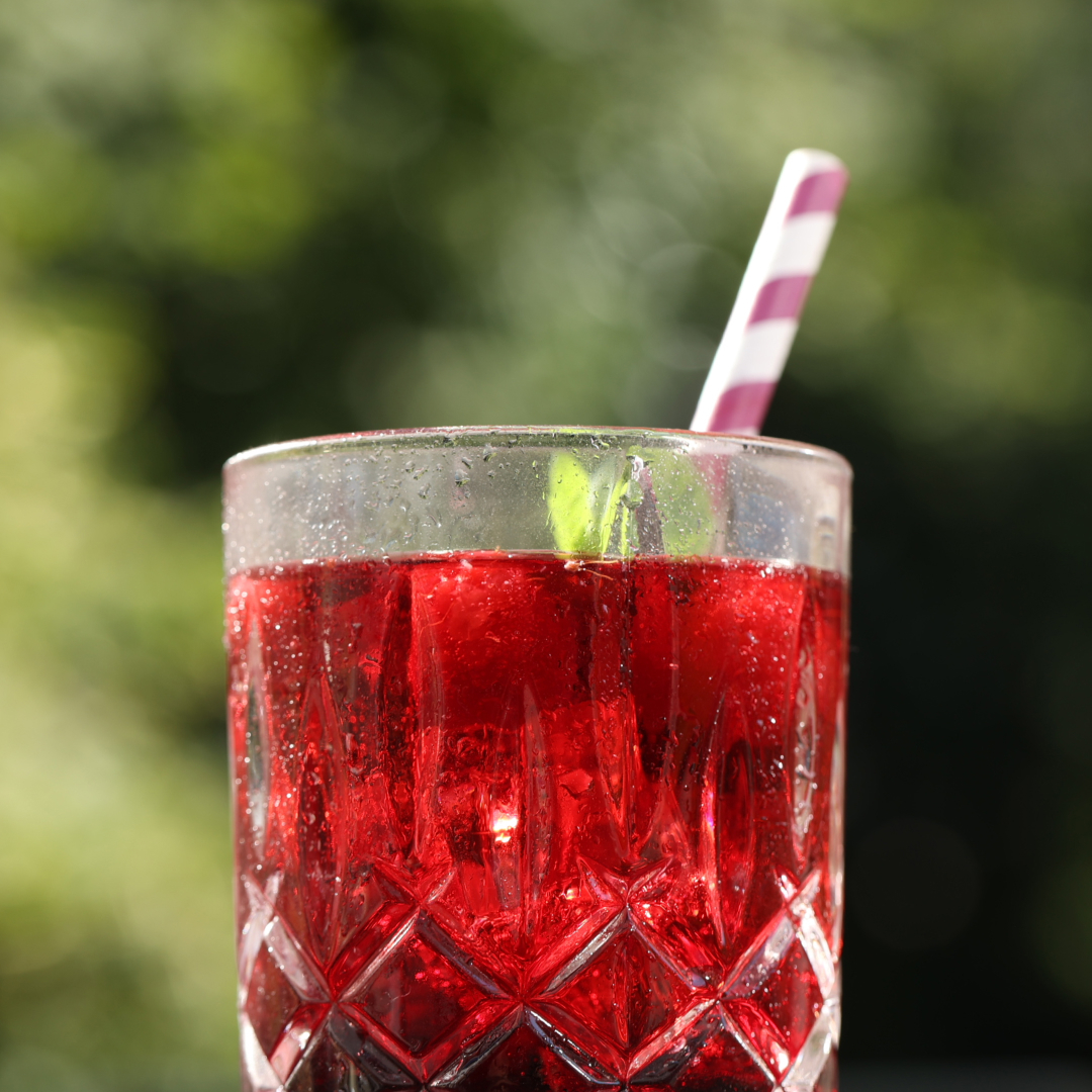 Glas mit roter Flüssigkeit, Eiswürfeln und gestreiftem Strohhalm. Grüner, verschwommener Hintergrund.