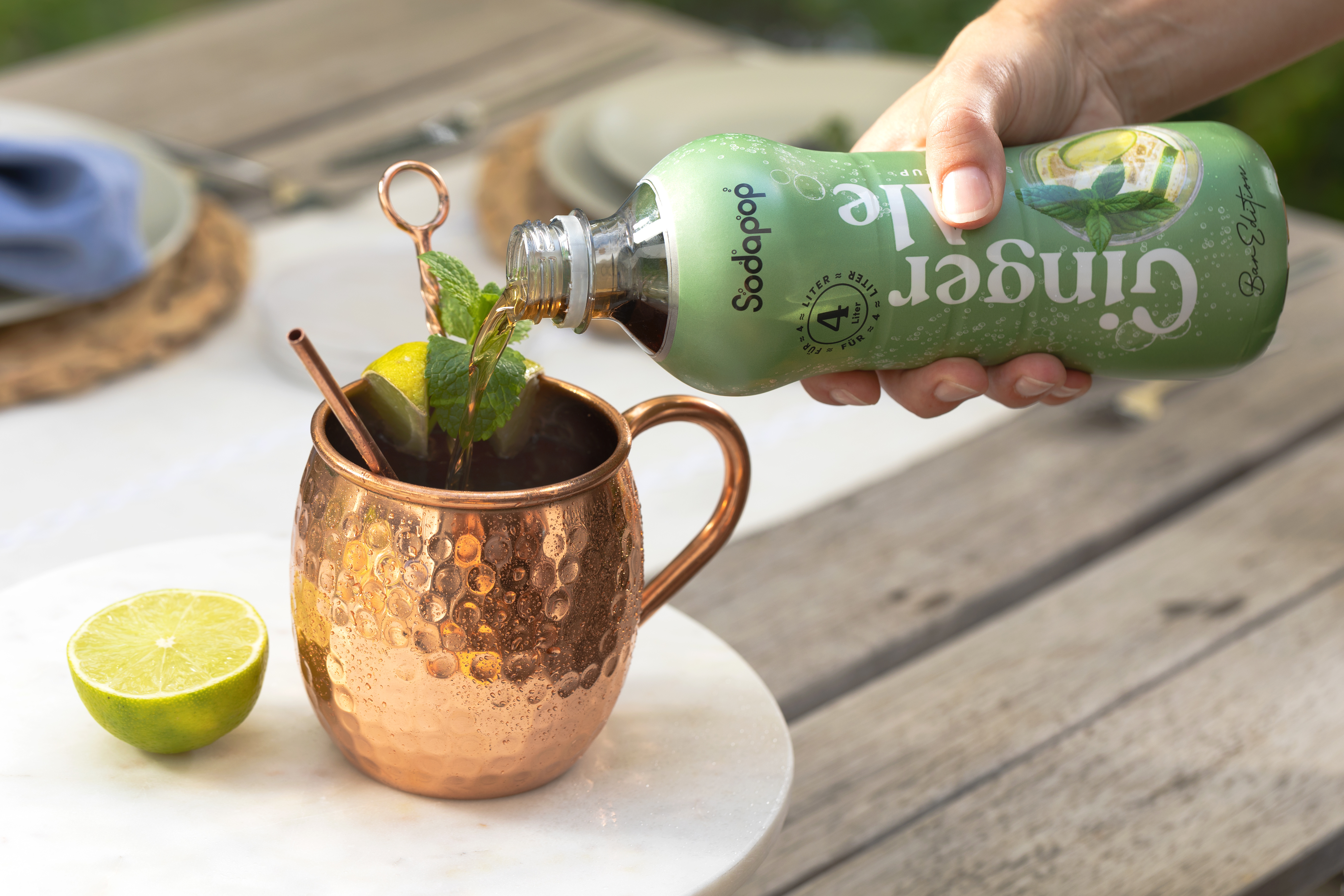 Hand gießt Ginger Ale in einen Kupferbecher mit Minze und Limette. Umgebung im Freien.