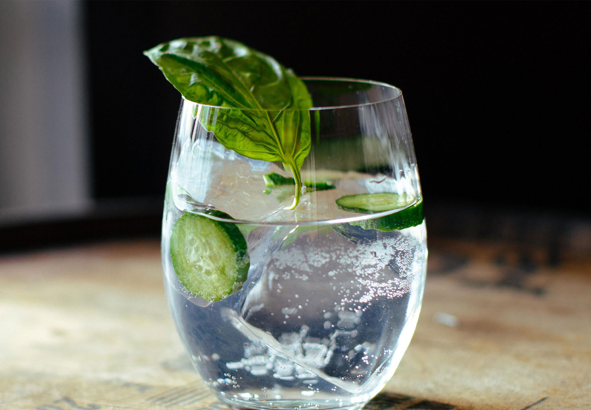 Ein Cocktailglas mit Eis, Gurkenscheiben, Basilikum und Sprudelwasser auf einer Holzoberfläche.