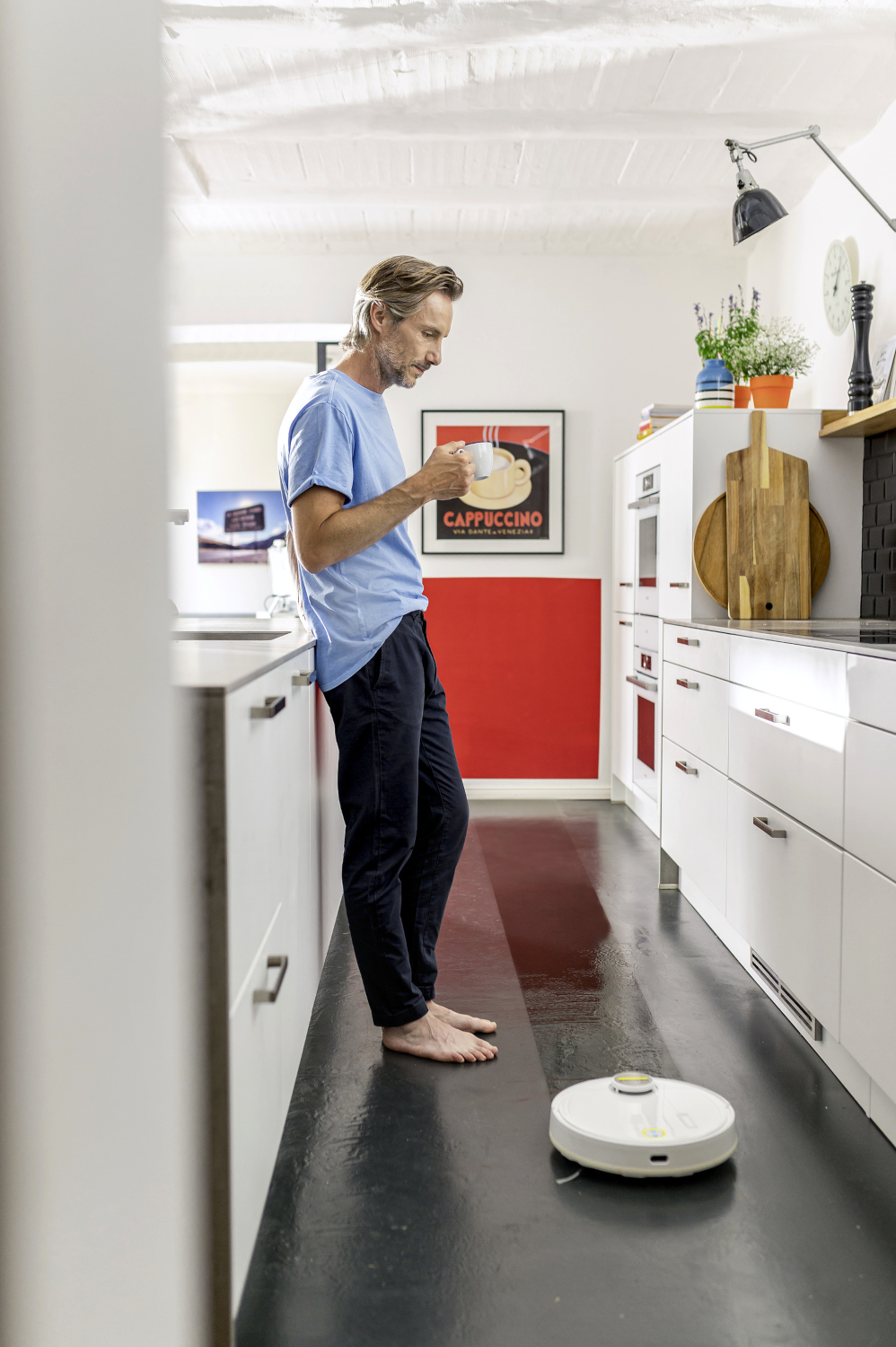 Egy kék inget viselő férfi egy konyhai pultra támaszkodik, egy robotporszívó takarítja a padlót.