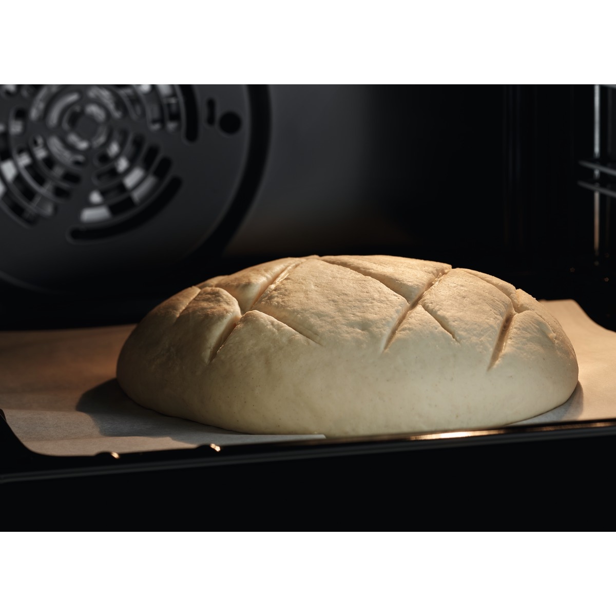 Bochenek chleba, nacięty na wierzchu, leży na papierze do pieczenia wewnątrz ciemnego piekarnika.