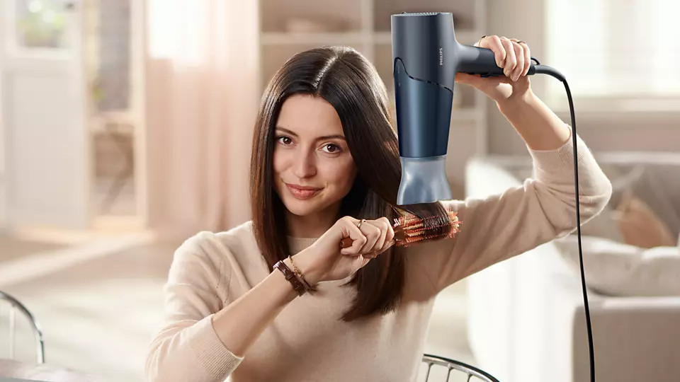 Woman brushing her hair with a brush while using a hairdryer, smiling.
