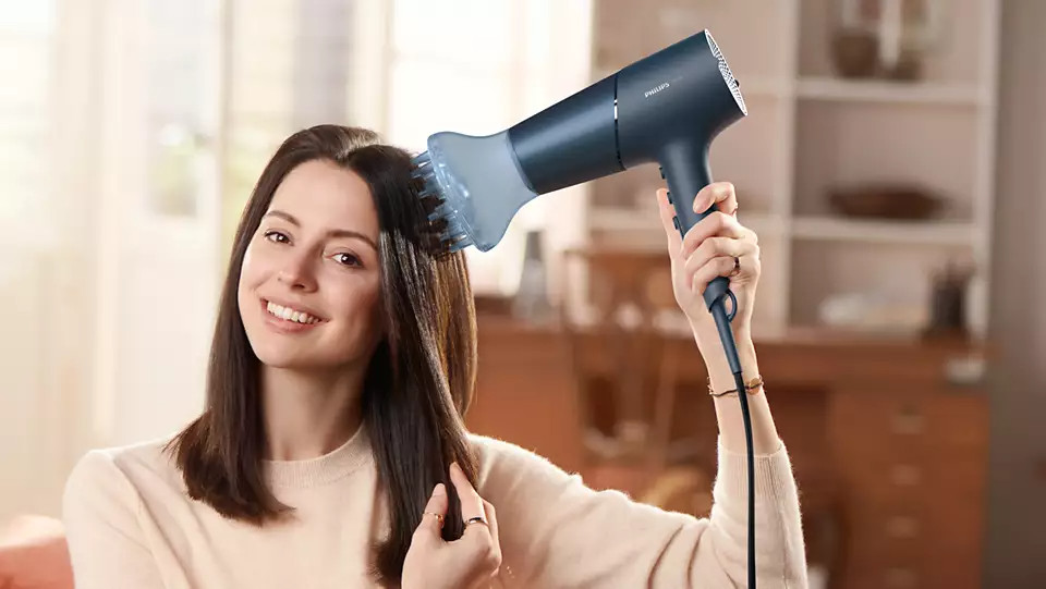 Woman using a dark blue hair dryer on her dark hair, smiling.