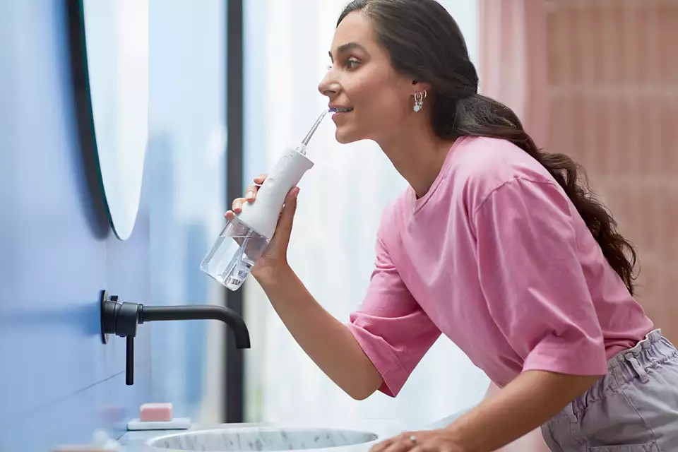 A woman using a water flosser in a bathroom, reflected in a mirror.