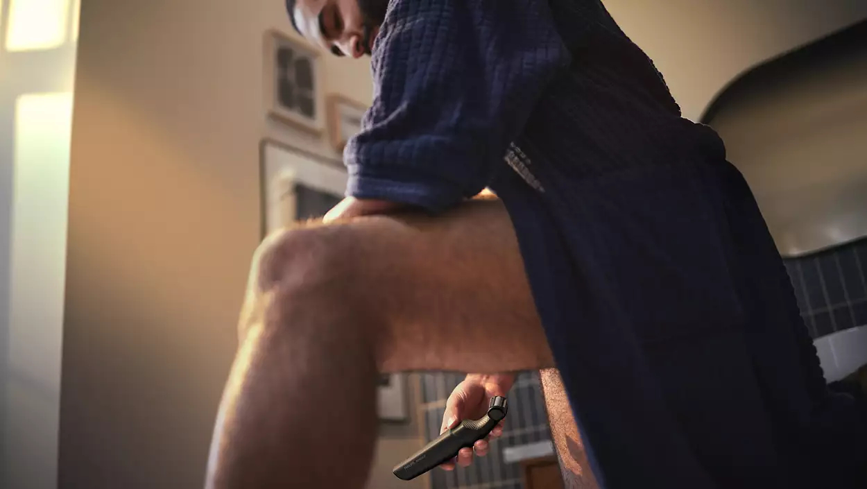 Mężczyzna goli nogę trymerem w łazience, ujęcie z niskiego kąta.