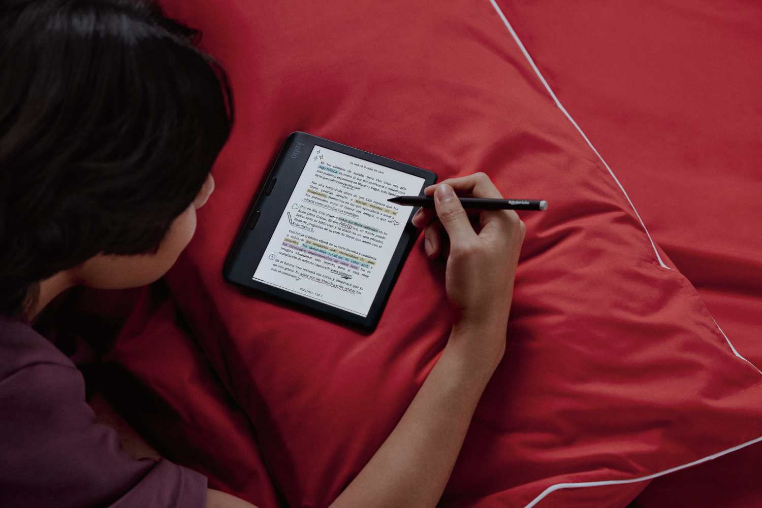 Persona leyendo en un lector Kobo, usando un lápiz. Fondo de almohada roja.