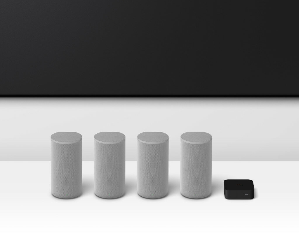 Cztery białe głośniki i czarne urządzenie na białym stole. Czarny telewizor w tle.